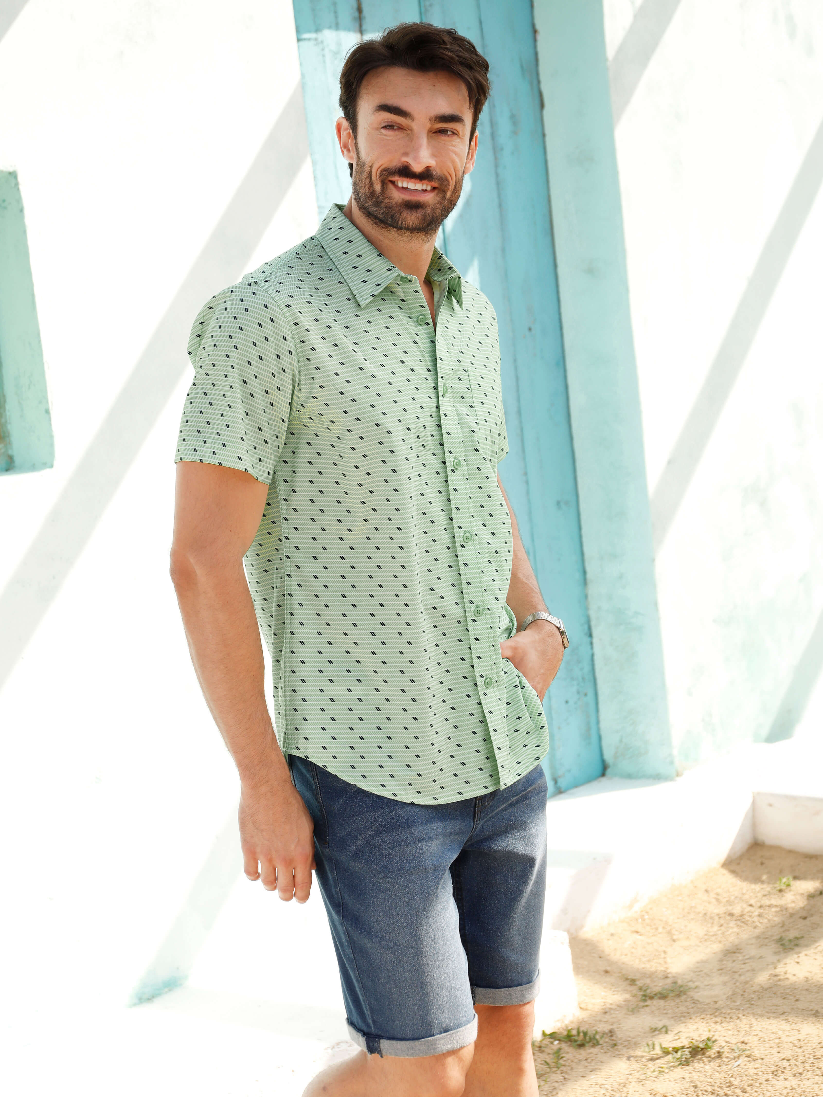 Ein männliches Model trägt ein kurzärmeliges, mintgrünes Hemd mit Muster und dunkelblaue Jeansshorts, steht an einer weißen Wand mit türkisfarbenen Akzenten.