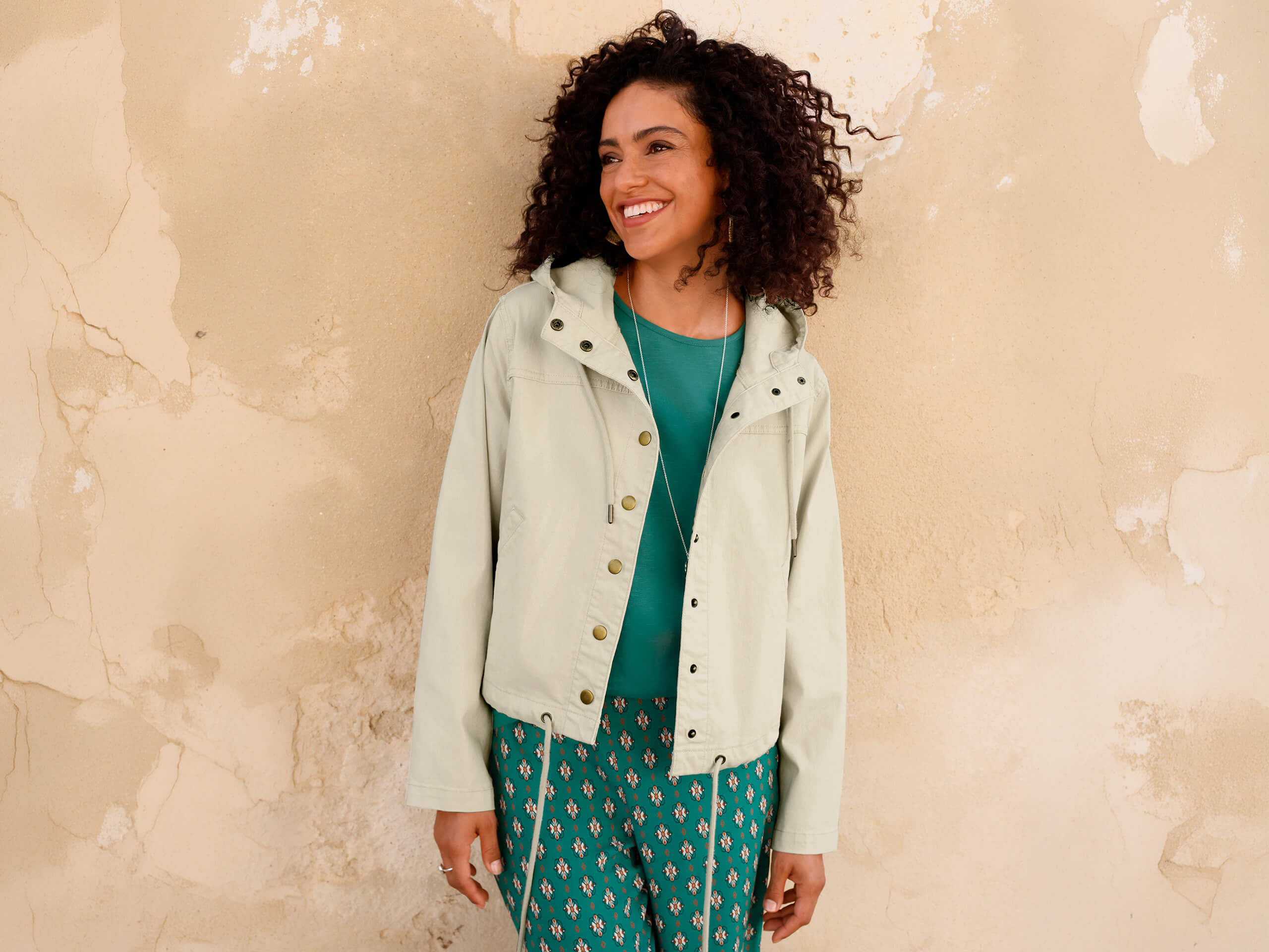 Ein Model trägt eine helle Jacke mit Kapuze über einem grünen Shirt und gemusterter grüner Hose, steht vor einer beigen Wand.