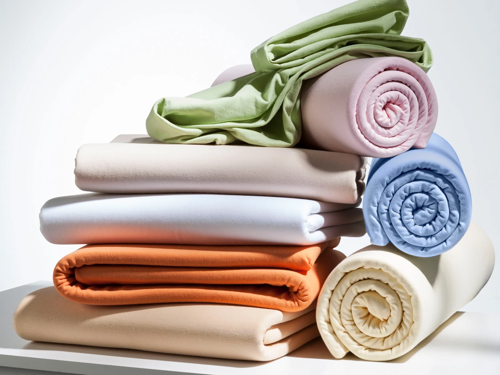 Stapel gefaltener und gerollter Stoffe in verschiedenen Farben wie Grün, Rosa, Blau, Orange und Beige auf weißer Oberfläche.