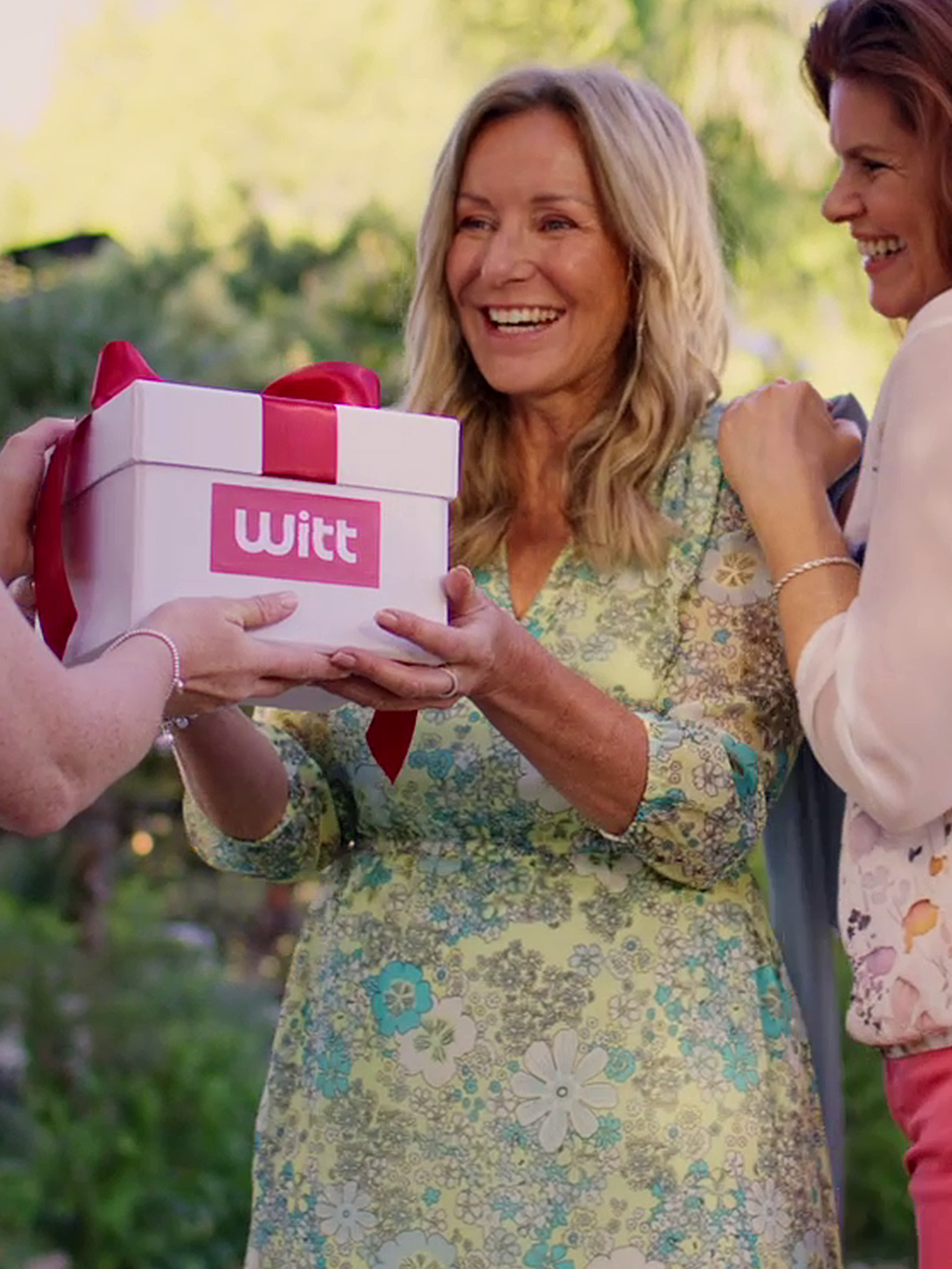 Zwei Frauen in floralen Kleidern im Freien. Die Frau mit lockigem Haar überreicht einer lachenden Frau eine weiße Geschenkbox mit dem 'Witt'-Logo und roter Schleife.