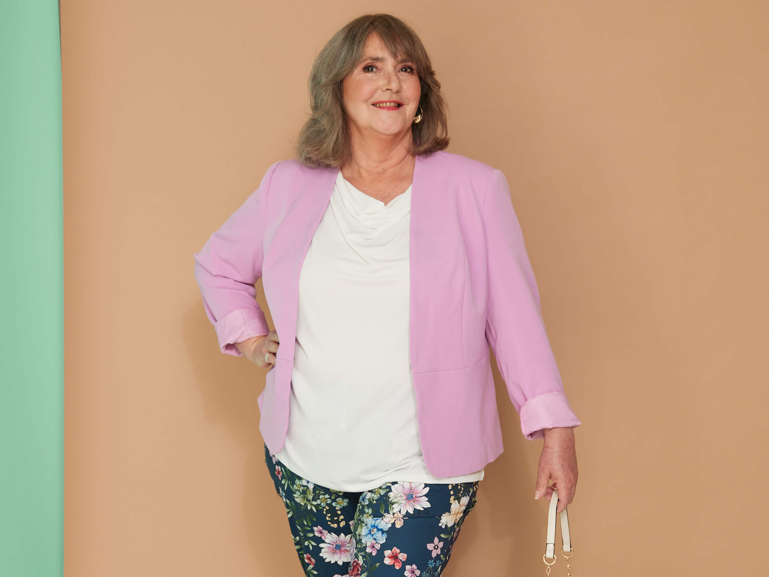 Ein Model trägt einen rosa Blazer, ein weißes Shirt und eine geblümte Hose vor braun-grünem Hintergrund.