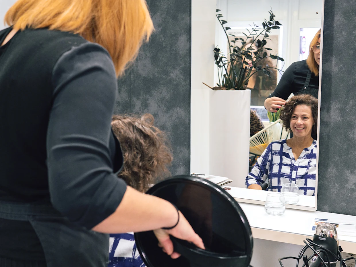 Eine Frau in weiß blau karierter Bluse mit kurzen braunen Haaren sitzt vor einem Spiegel. Hinter ihr steht eine rothaarige Frau in schwarzem Oberteil mit einem runden Friseurspiegel in der Hand. Eine Frau in weiß blau karierter Bluse mit kurzen braunen Haaren sitzt vor einem Spiegel. Hinter ihr steht eine rothaarige Frau in schwarzem Oberteil mit einem runden Friseurspiegel in der Hand.