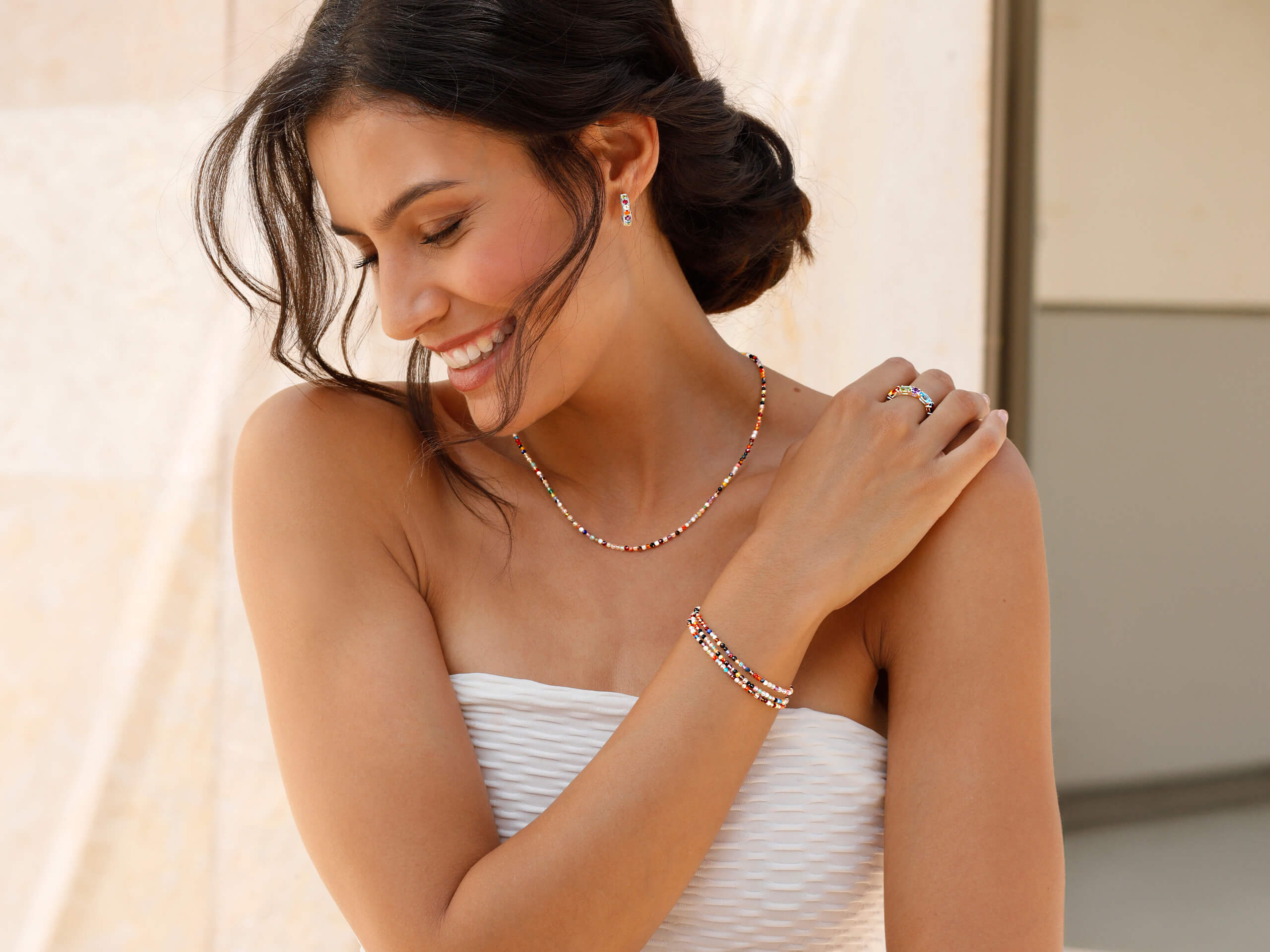Ein Model trägt ein weißes, ärmelloses Oberteil und bunten Schmuck aus Perlen an Hals, Handgelenk und Finger, steht vor hellem Hintergrund.