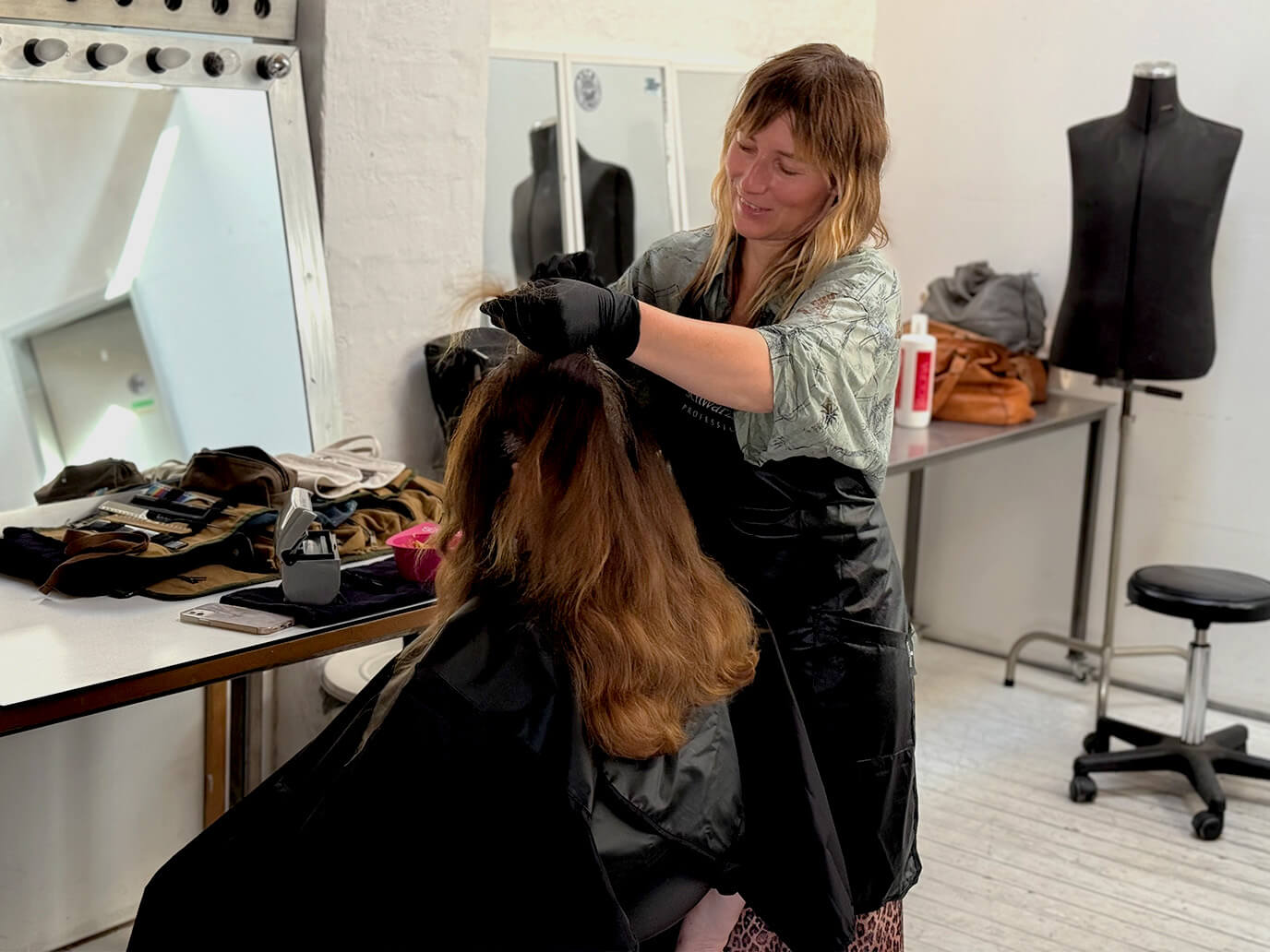 Eine Frau sitzt mit einem schwarzen Umhang vor einem Spiegel, während eine Friseurin mit Handschuhen das Haar der Frau hochhebt.