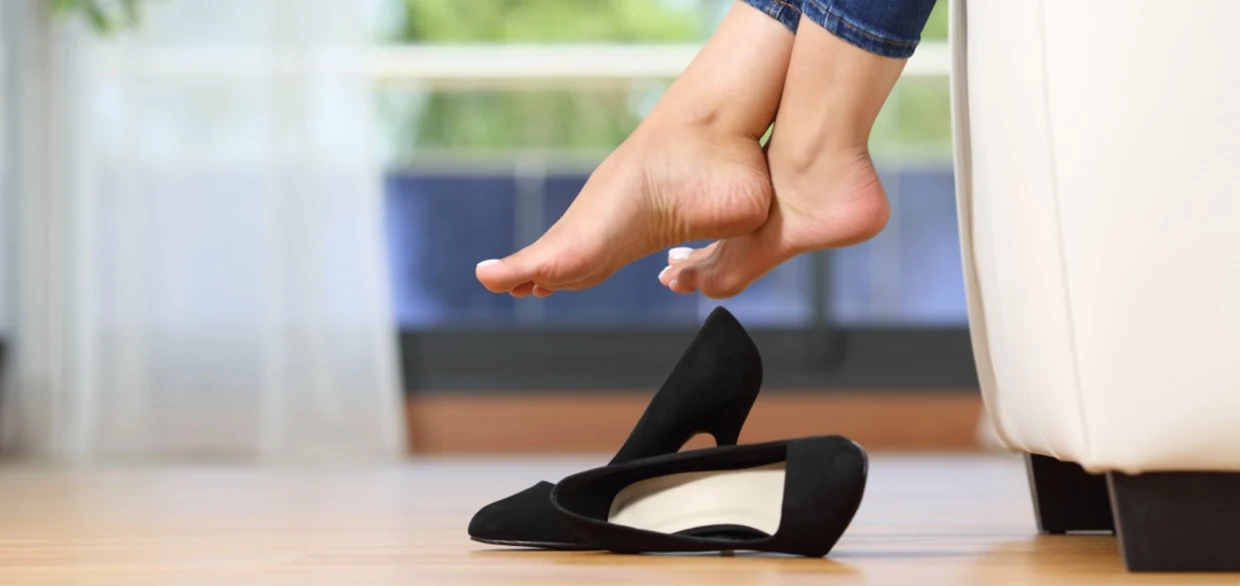 Barfuße Füße einer Person ragen von einem Sofa herab, schwarze High-Heels stehen auf dem Holzboden daneben. Barfuße Füße einer Person ragen von einem Sofa herab, schwarze High-Heels stehen auf dem Holzboden daneben.