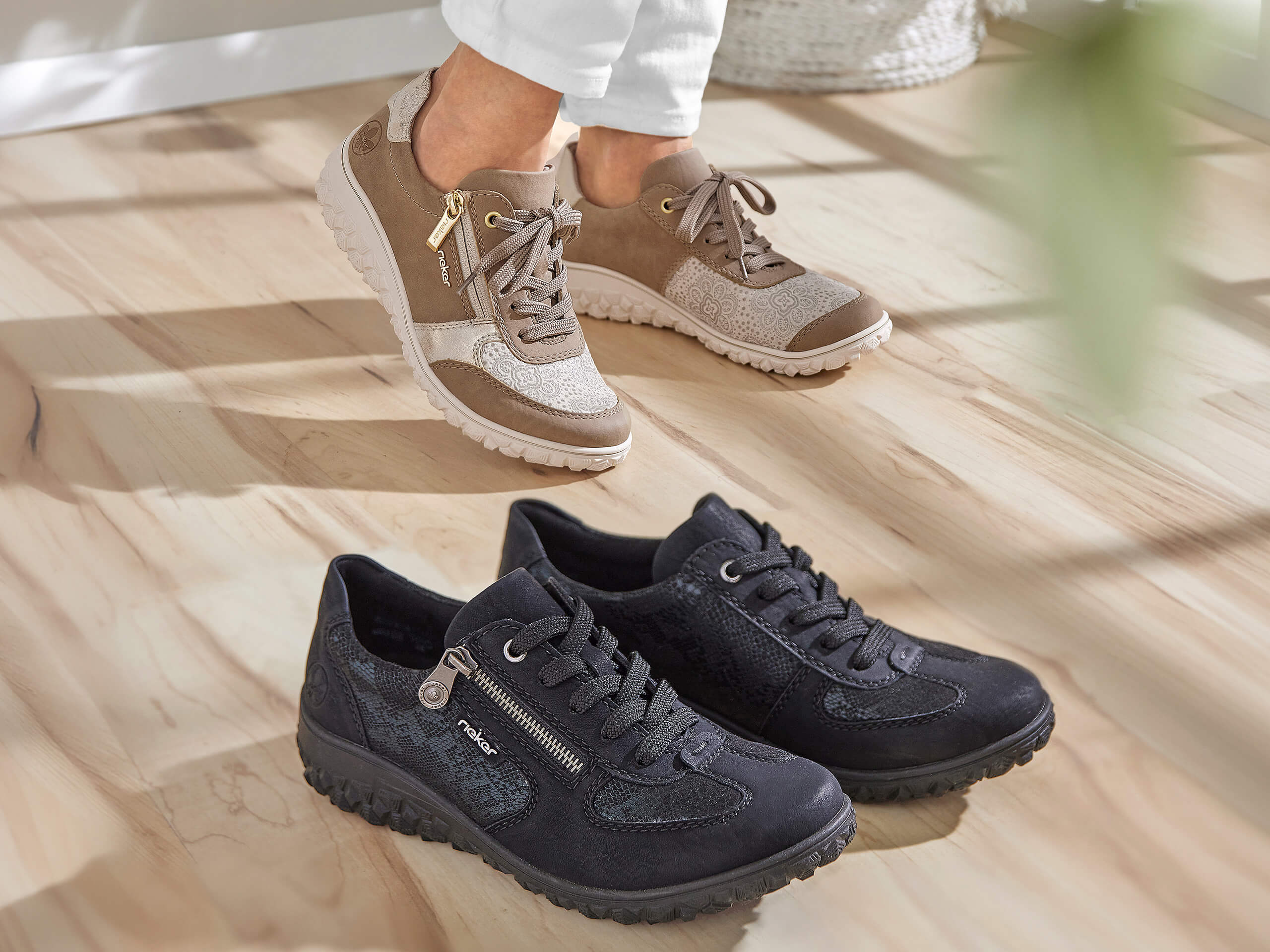 Eine Frau trägt ein Paar beige Sneaker, daneben steht ein Paar schwarze Sneaker auf einem hellen Holzboden.