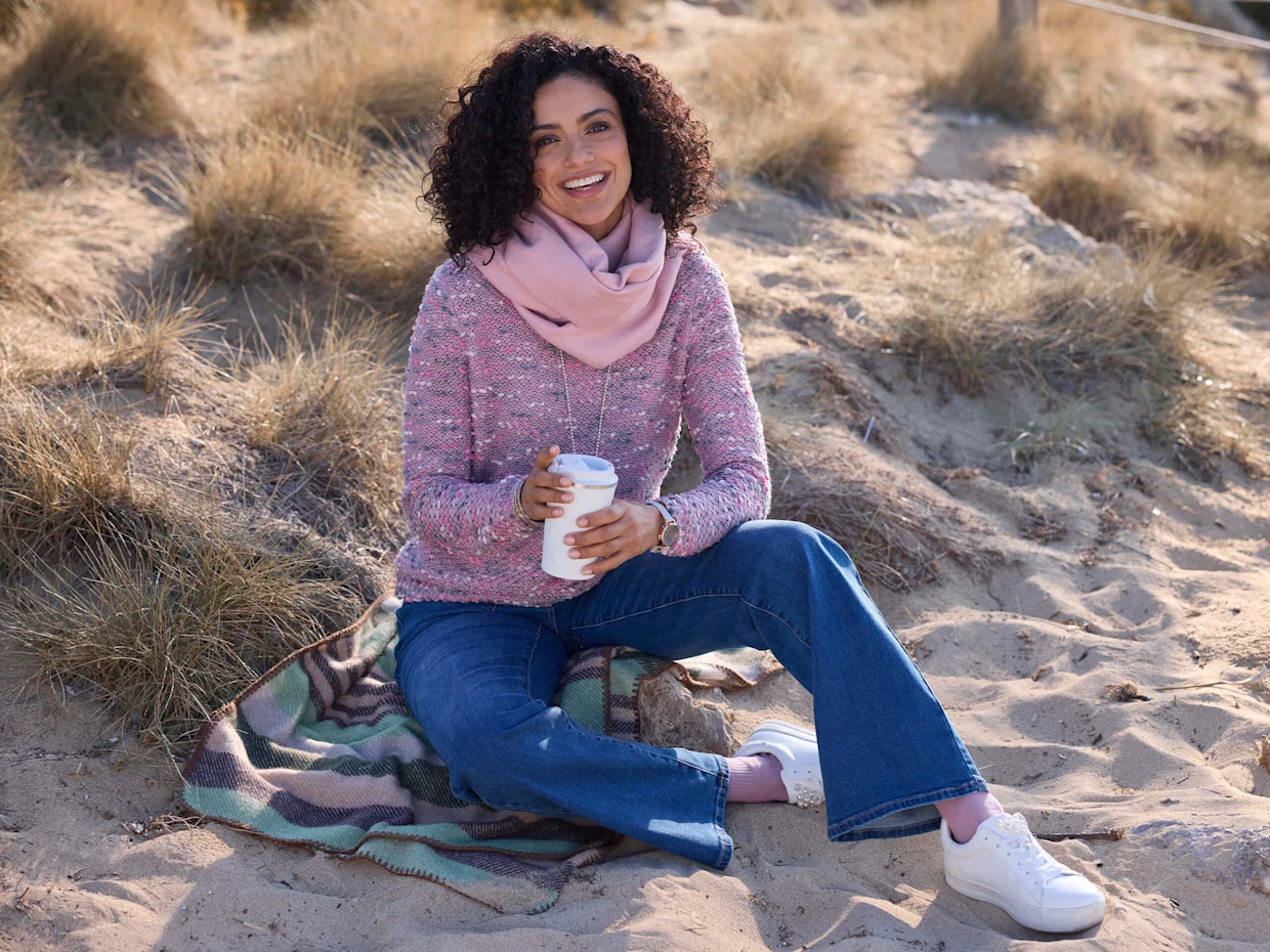 Ein Model sitzt auf einer Decke im Sand, trägt einen rosa Pullover, einen passenden Schal, blaue Jeans und weiße Sneaker. Sie hält einen Becher.