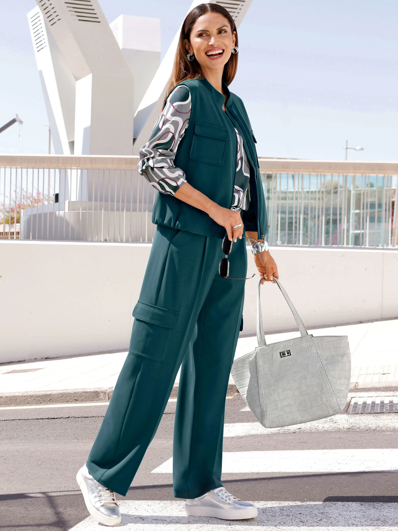 Ein Model trägt ein petrolfarbenes Outfit mit gemusterter Bluse, Sneakern und heller Handtasche im Freien. Ein Model trägt ein petrolfarbenes Outfit mit gemusterter Bluse, Sneakern und heller Handtasche im Freien.