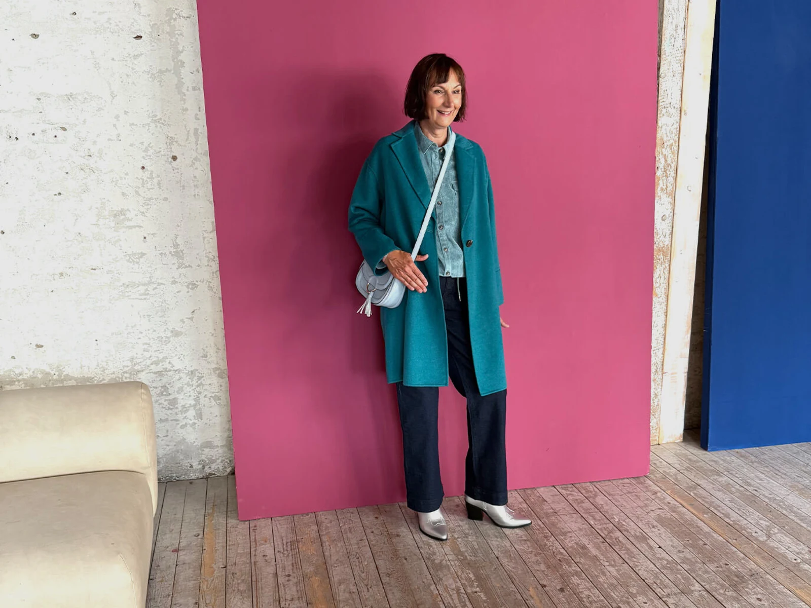 Eine Frau trägt einen türkisfarbenen Mantel, eine hellblaue Jeansjacke, dunkle Hosen und silberne Stiefeletten vor einer pinken Wand. Eine Frau trägt einen türkisfarbenen Mantel, eine hellblaue Jeansjacke, dunkle Hosen und silberne Stiefeletten vor einer pinken Wand.