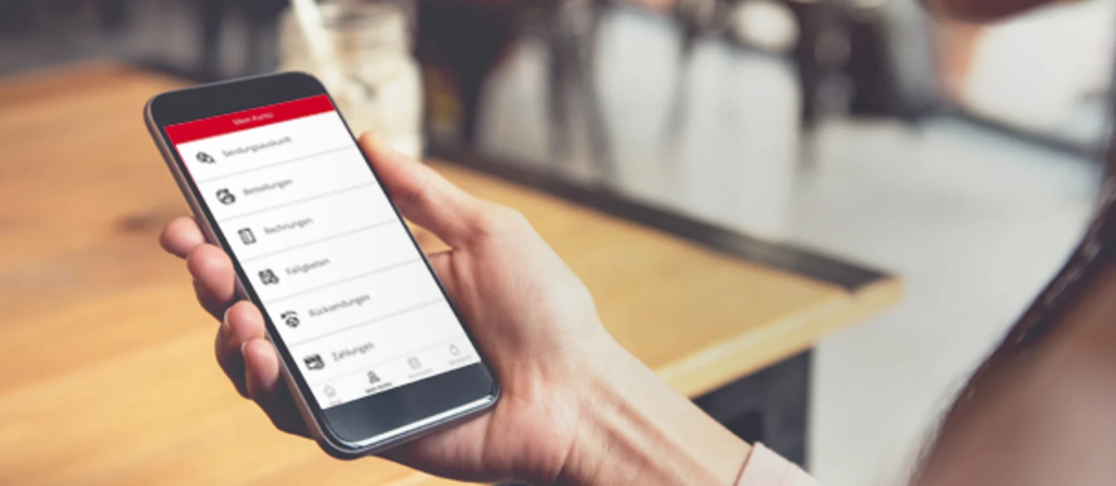 Person hält ein Smartphone mit geöffneter App in der Hand, im Hintergrund ein Tisch in einem Café. Person hält ein Smartphone mit geöffneter App in der Hand, im Hintergrund ein Tisch in einem Café.