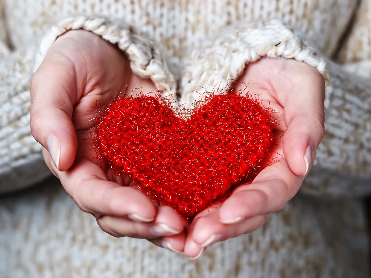 Hände in einem Wollpullover halten ein rotes, glitzerndes Herz aus Stoff.