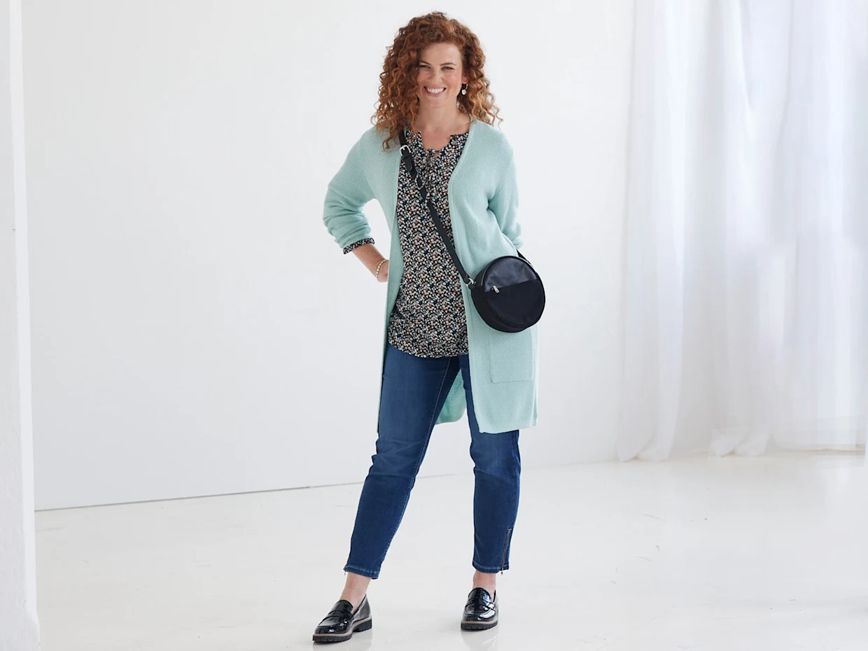 Frau trägt ein mintgrünes Strick-Cardigan, gemustertes Oberteil, blaue Jeans und schwarze Loafer; schwarze Umhängetasche rundet das Outfit ab.