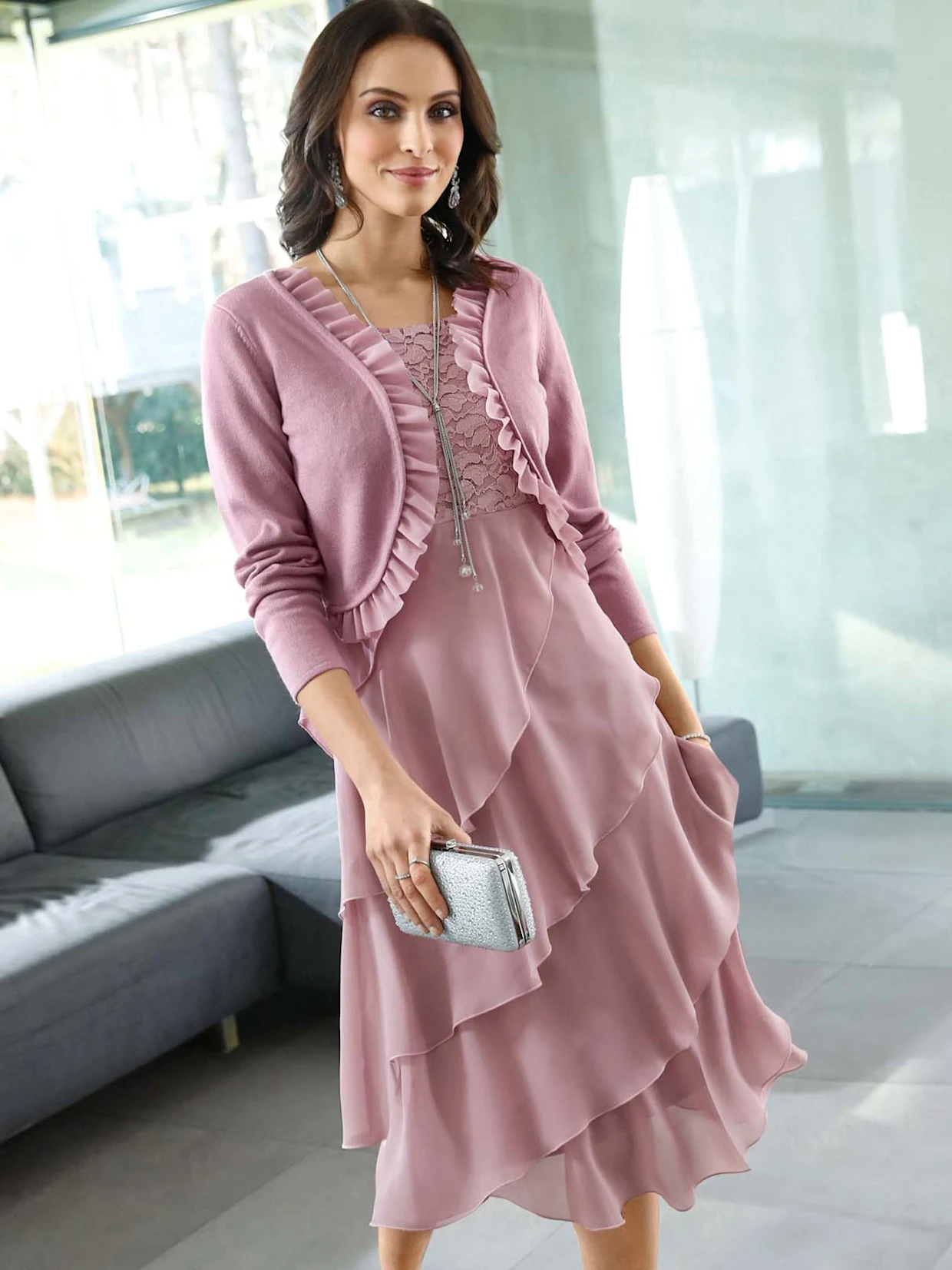 Frau trägt ein elegantes Outfit in Rosa, bestehend aus einer gerüschten Strickjacke und einem mehrlagigen Chiffonkleid, kombiniert mit einer silbernen Clutch. Hintergrund ist ein modernes Wohnzimmer mit großen Glasfenstern.