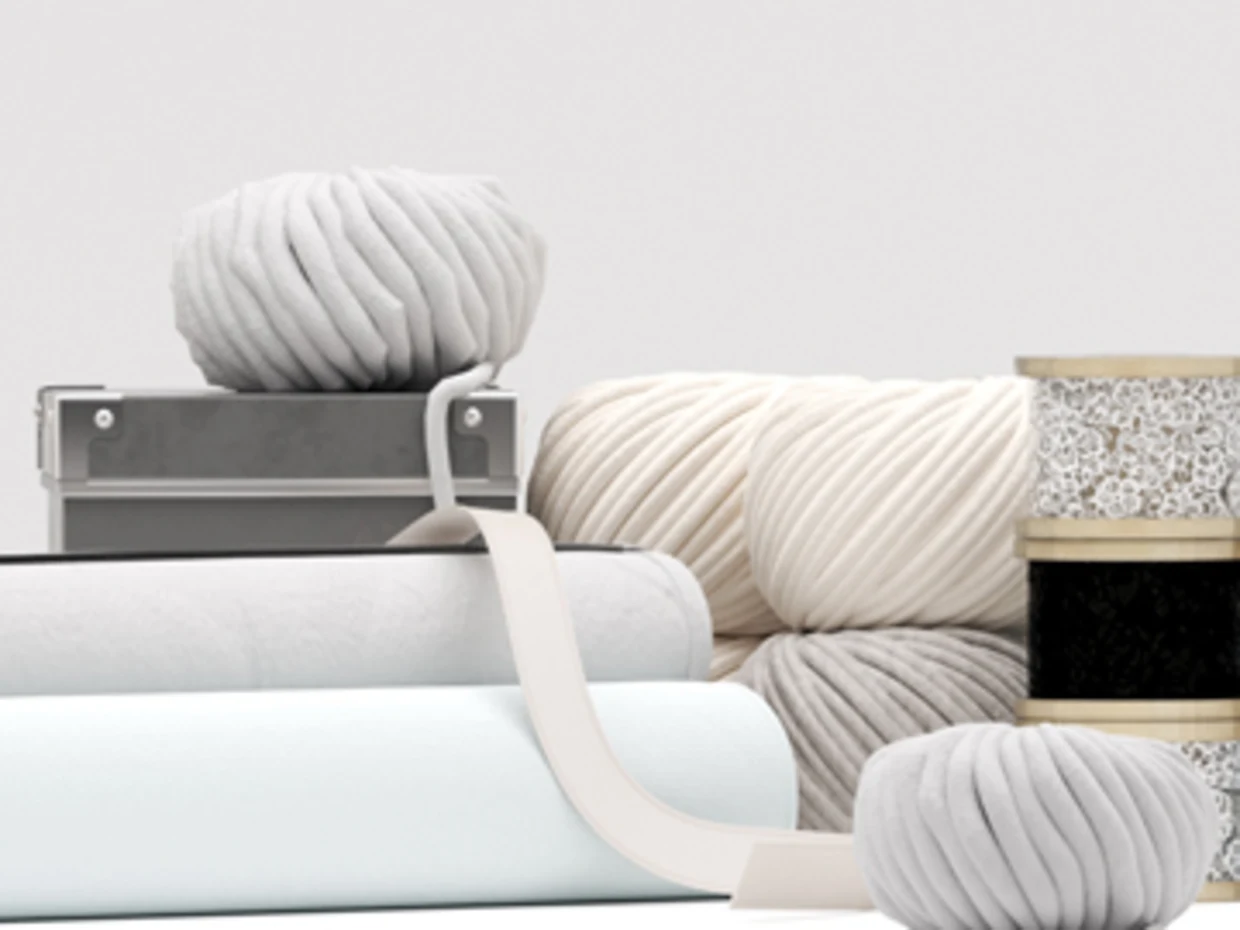 Wollknäuel in Grautönen, Stoffrollen und Spitzenbänder vor neutralem Hintergrund.