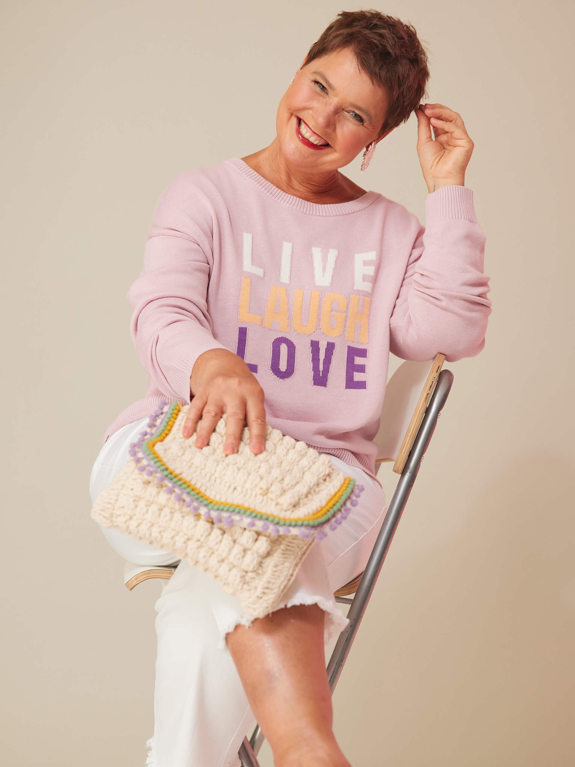 Eine Frau mit kurzen braunen Haaren trägt einen rosa Pullover mit der Aufschrift 'LIVE LAUGH LOVE', weiße Hosen und hält eine gehäkelte Tasche, sitzt auf einem Stuhl und lächelt.