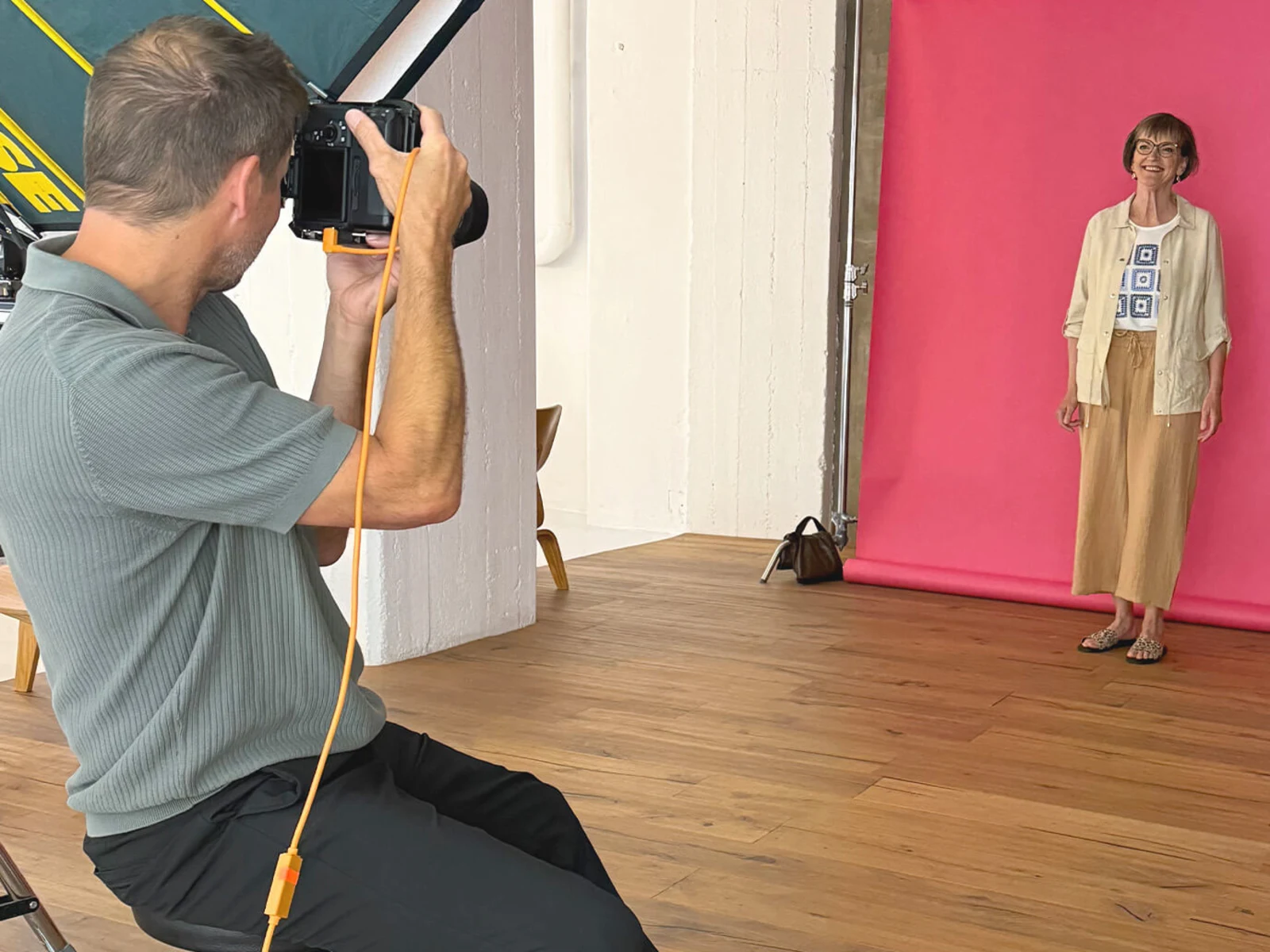 Ein Fotograf sitzt und macht ein Foto von einer Frau, die vor einem pinken Hintergrund steht. Die Frau trägt eine helle Jacke und eine weite Hose. Ein Fotograf sitzt und macht ein Foto von einer Frau, die vor einem pinken Hintergrund steht. Die Frau trägt eine helle Jacke und eine weite Hose.
