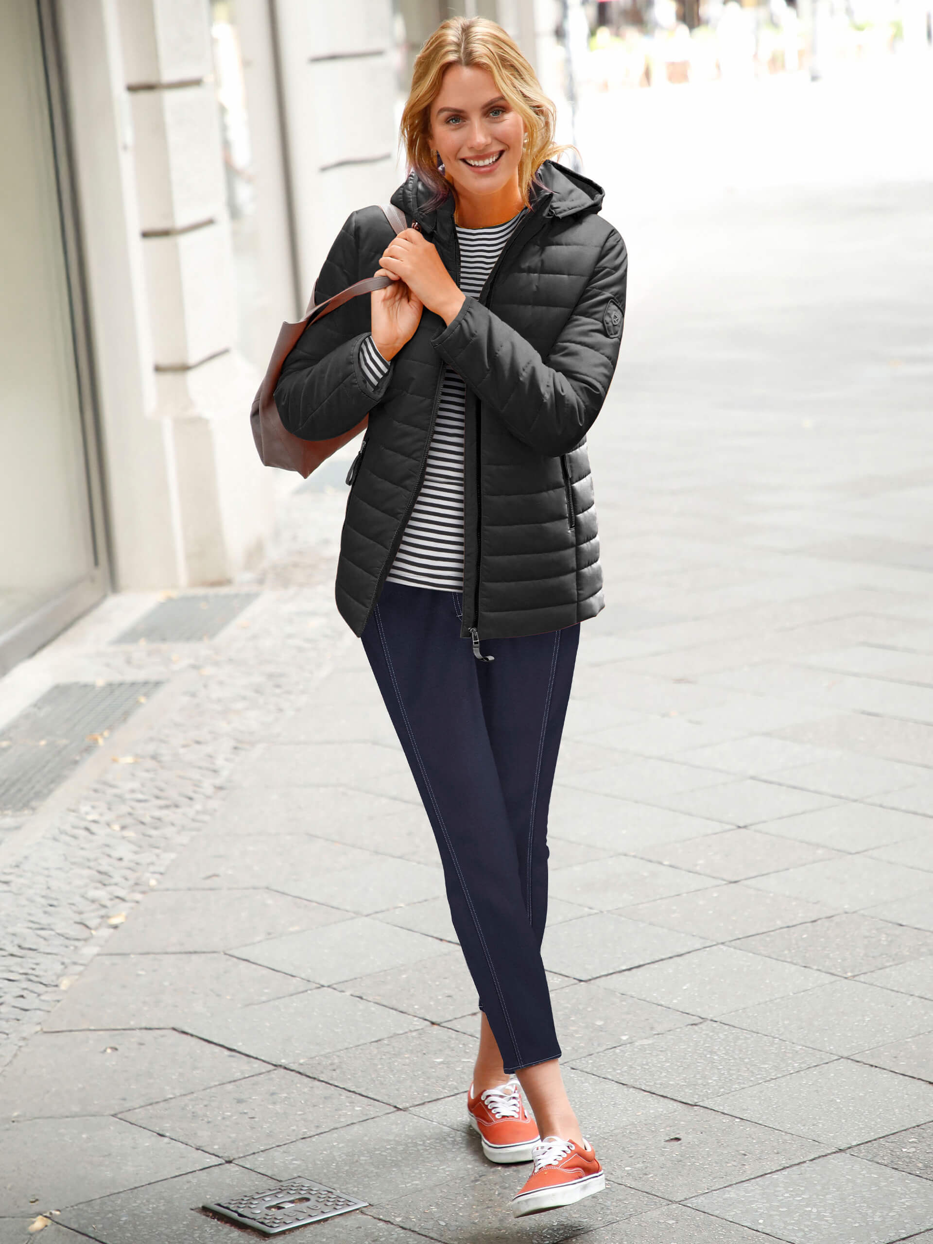 Ein Model geht auf der Straße, trägt eine schwarze Jacke, gestreiftes Oberteil und dunkelblaue Hosen, mit Sneakers.