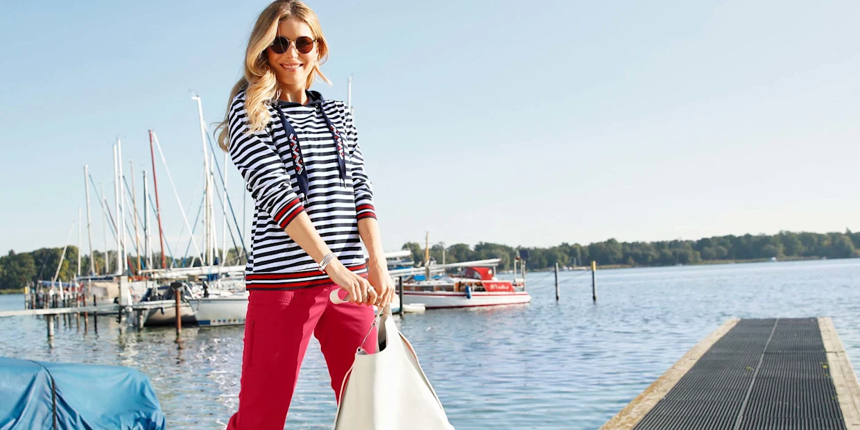 Frau in maritim gestreiftem Pullover und roter Hose steht mit weißer Tasche auf einem Steg vor einem Hafen mit Segelbooten. Frau in maritim gestreiftem Pullover und roter Hose steht mit weißer Tasche auf einem Steg vor einem Hafen mit Segelbooten.