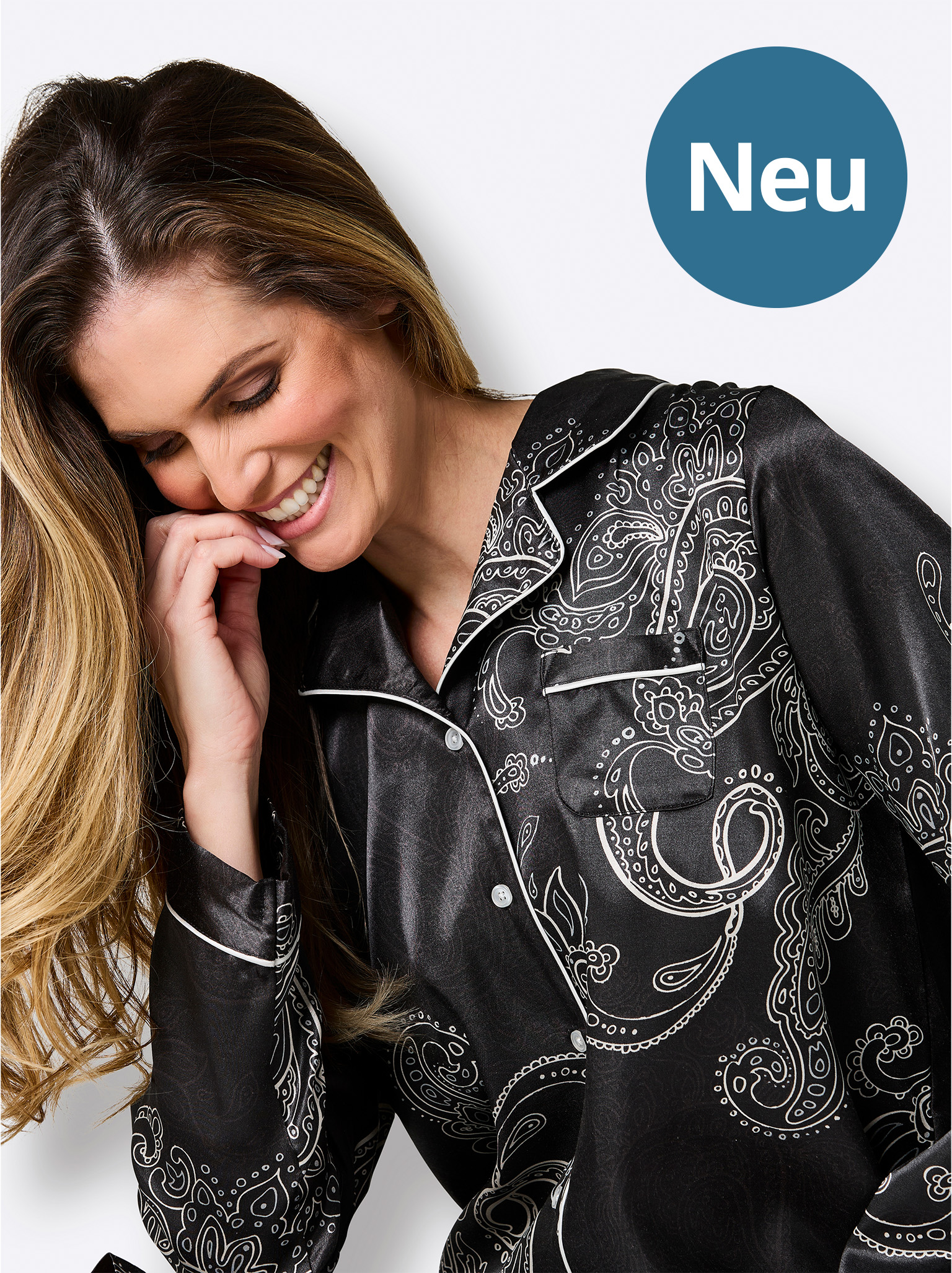 Lächelnde Frau im schwarzen Paisley-Schlafanzug blickt nach unten, neben blauem Kreis mit „Neu“.