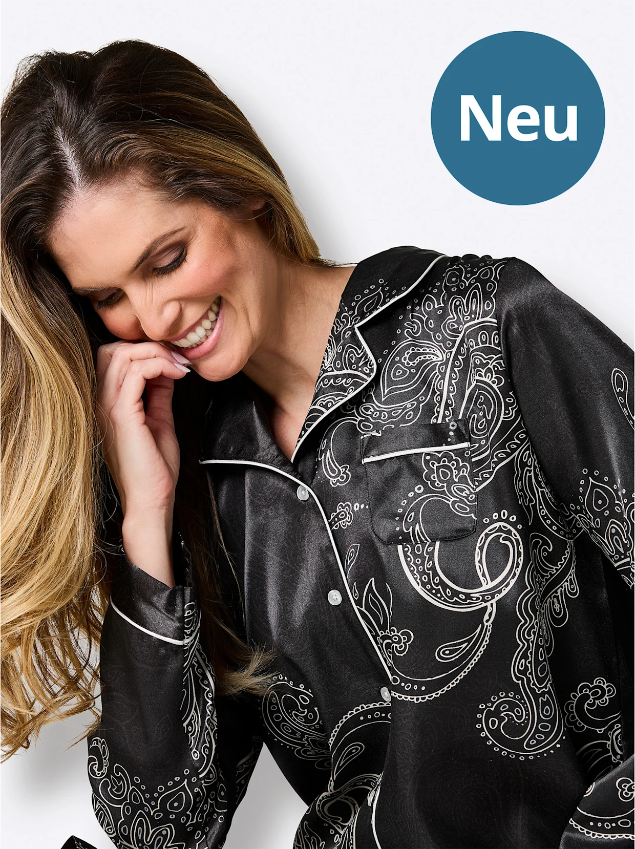 Lächelnde Frau im schwarzen Paisley-Schlafanzug blickt nach unten, neben blauem Kreis mit „Neu“.