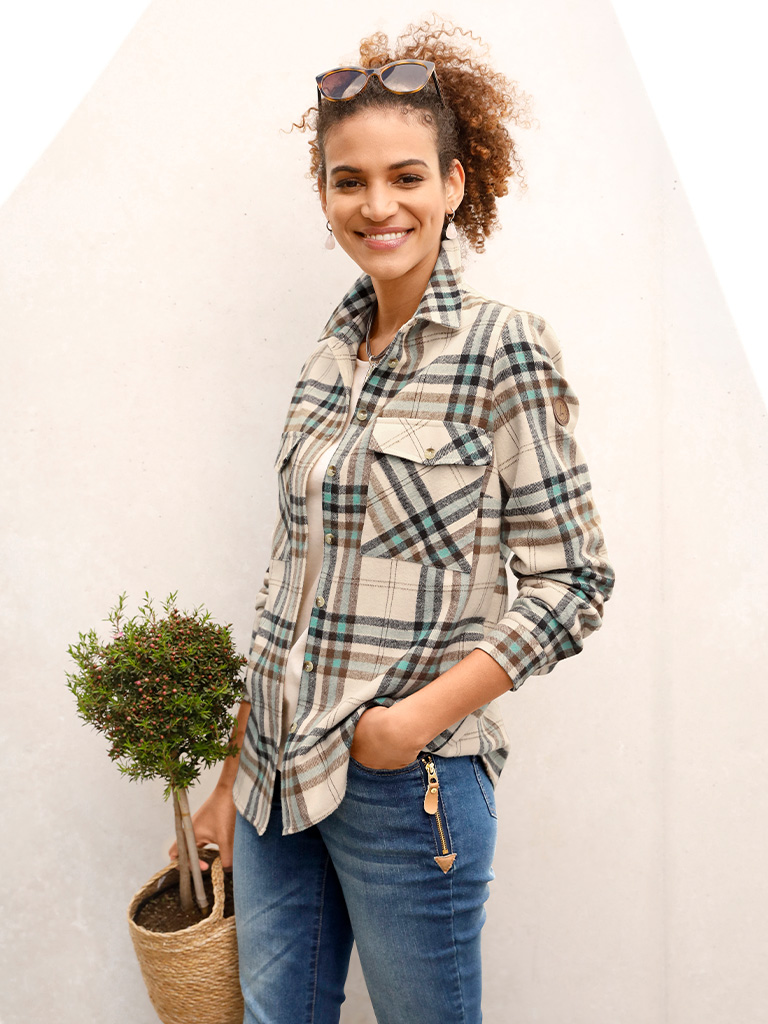 Ein Model trägt eine jade-elfenbeinfarbene Flanellbluse mit aufgekrempelten Ärmeln und blaue Jeans, hält einen kleinen Topf mit Pflanze.