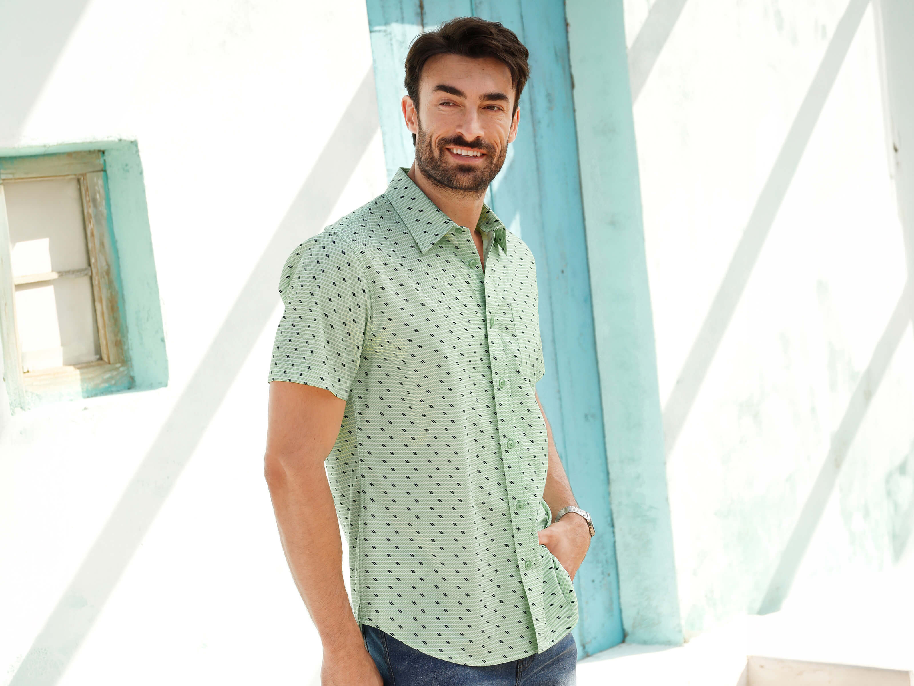 Ein männliches Model trägt ein kurzärmeliges, mintgrünes Hemd mit Muster und dunkelblaue Jeansshorts, steht an einer weißen Wand mit türkisfarbenen Akzenten.