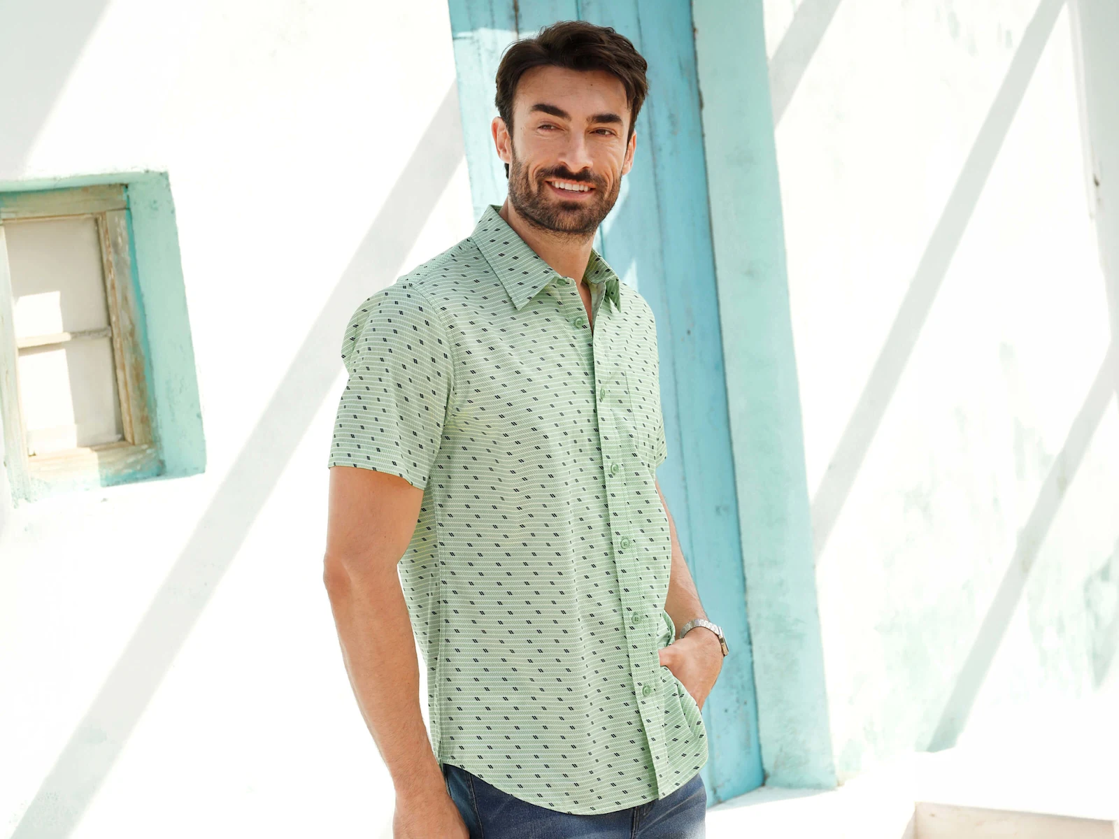 Ein männliches Model trägt ein kurzärmeliges, mintgrünes Hemd mit Muster und dunkelblaue Jeansshorts, steht an einer weißen Wand mit türkisfarbenen Akzenten.