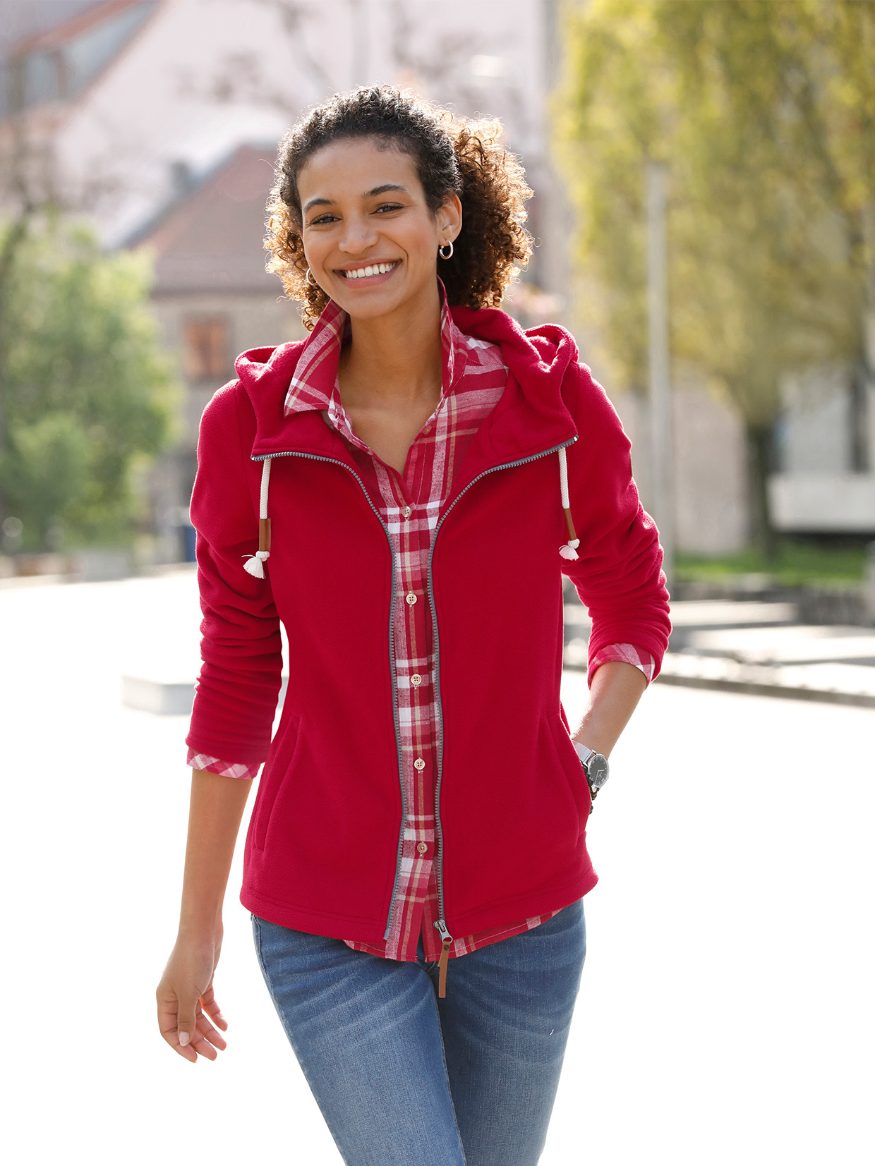 Frau trägt eine rote Fleecejacke mit Kapuze über einem karierten Hemd und kombiniert diese mit einer blauen Jeans, im Hintergrund sind Bäume und Gebäude zu sehen.