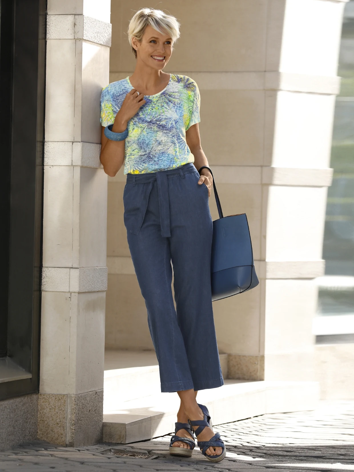 Frau trägt eine sommerliche Kombination aus gemustertem T-Shirt, blauer Stoffhose mit Gürtel und blauen Sandalen, dazu eine blaue Tasche; lässiger Look vor moderner Architektur.