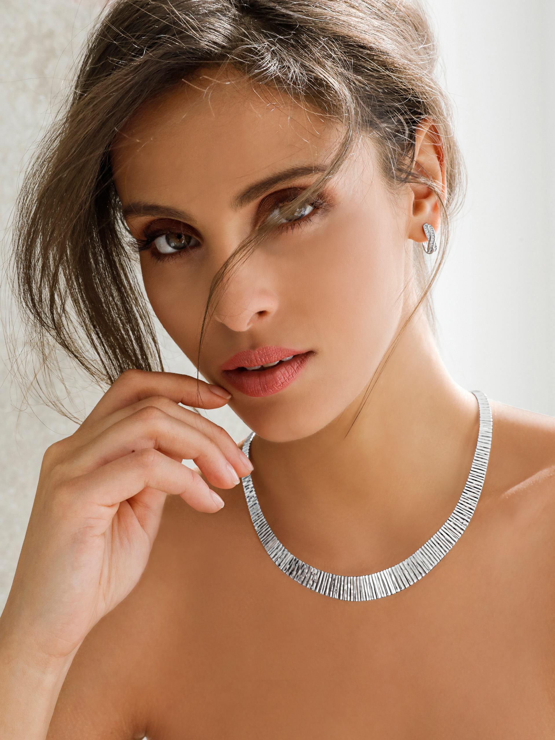 Ein Model trägt eine silberne Kette und passende Ohrringe, Haare sind locker zurückgebunden, Hintergrund ist hell.