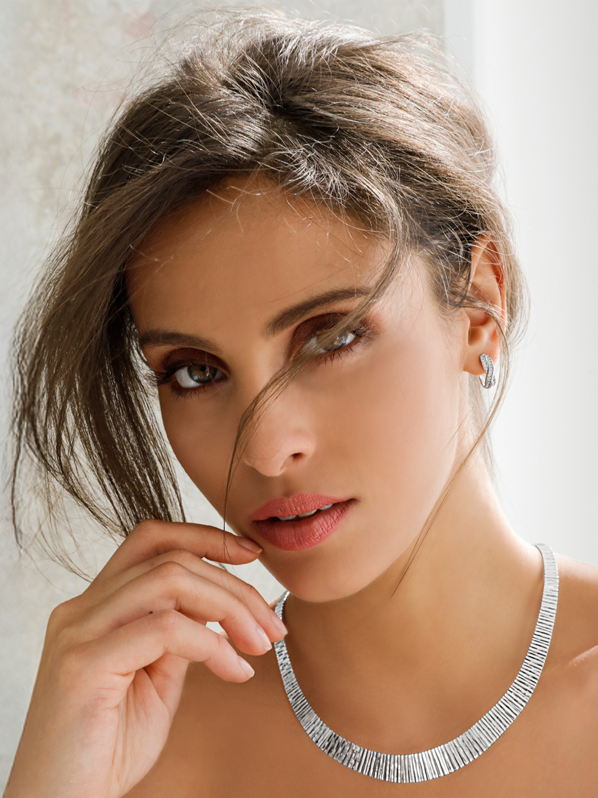 Een model draagt een zilveren ketting en bijpassende oorbellen, het haar is losjes naar achter gebonden, lichte achtergrond.