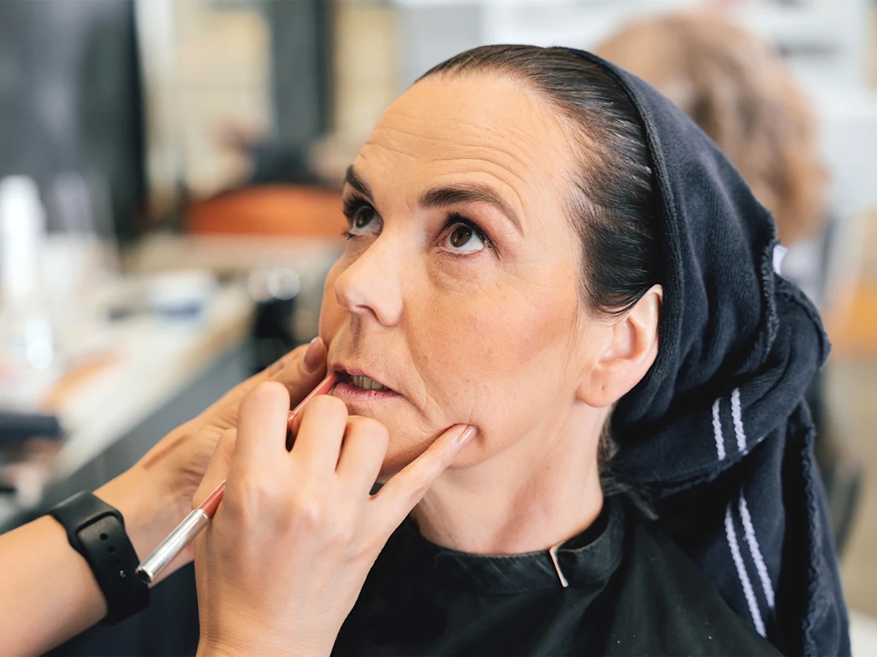 Eine dunkelhaarige Frau trägt einen Freiseurumhang und hat ein Handtuch um ihre nassen Haare gewickelt während sie gerade von einer anderen Person geschminkt wird.  Eine dunkelhaarige Frau trägt einen Freiseurumhang und hat ein Handtuch um ihre nassen Haare gewickelt während sie gerade von einer anderen Person geschminkt wird.