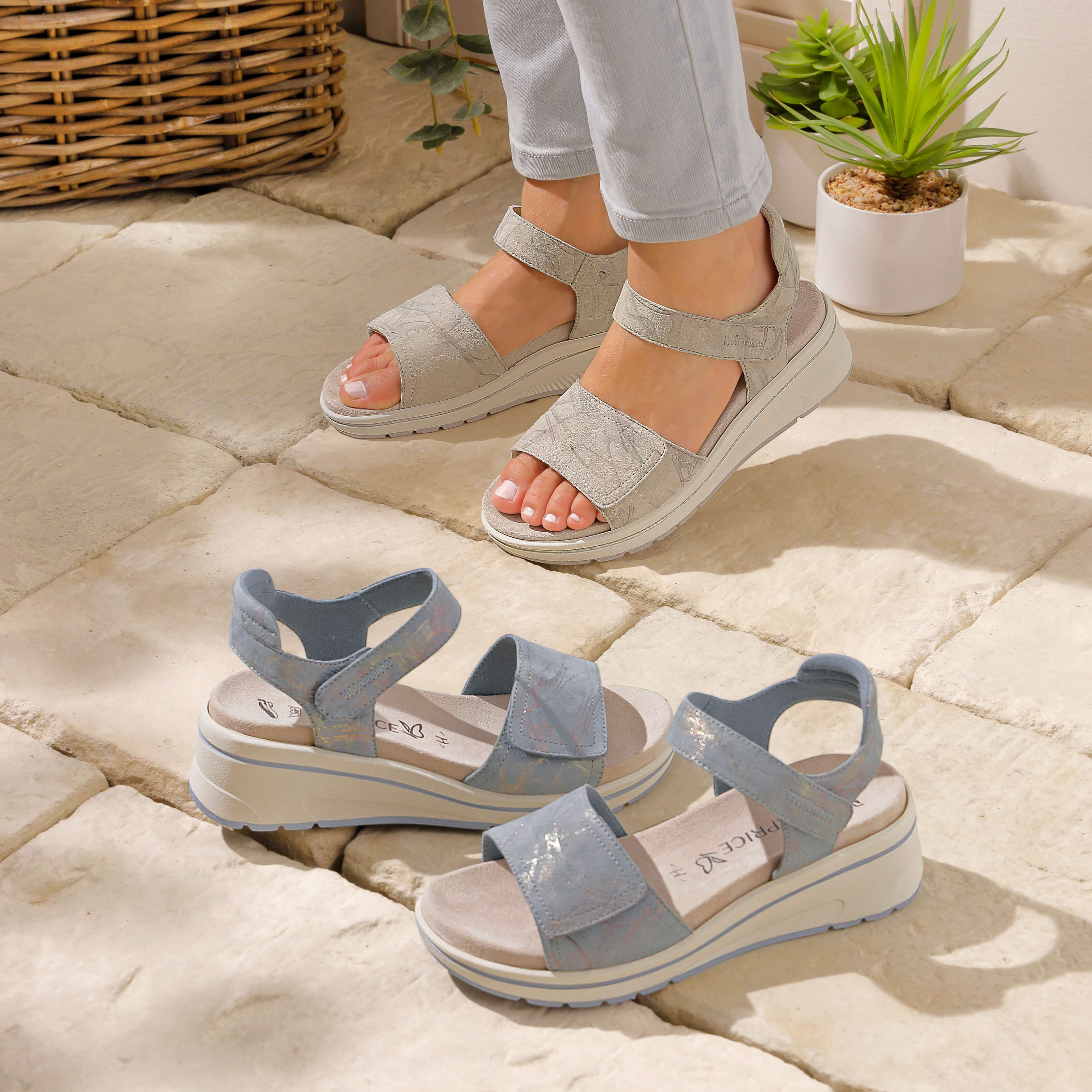 Ein Model trägt silberfarbene Sandalen mit Klettverschluss und Plateausohle, daneben stehen ähnliche Sandalen in Hellblau, Pflanzen im Hintergrund.