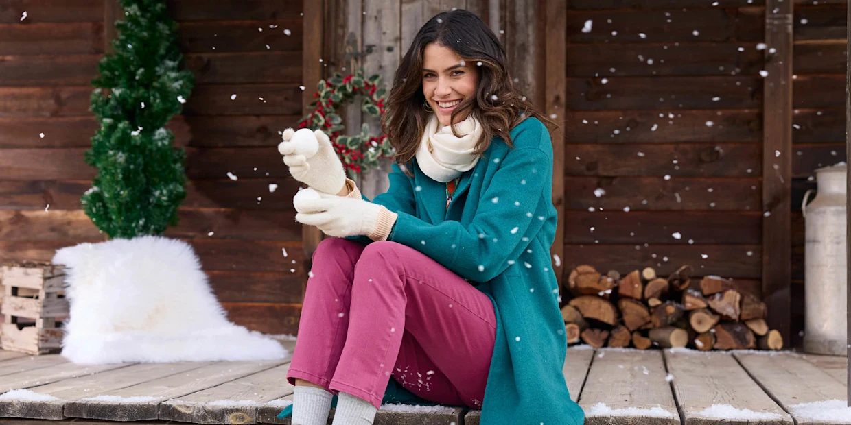 Ein Model in einem blauen Mantel, pinker Hose und braunen Stiefeletten sitzt auf einer Holzveranda, hält Schneebälle in den Händen, umgeben von Schnee und einer winterlichen Kulisse. Ein Model in einem blauen Mantel, pinker Hose und braunen Stiefeletten sitzt auf einer Holzveranda, hält Schneebälle in den Händen, umgeben von Schnee und einer winterlichen Kulisse.