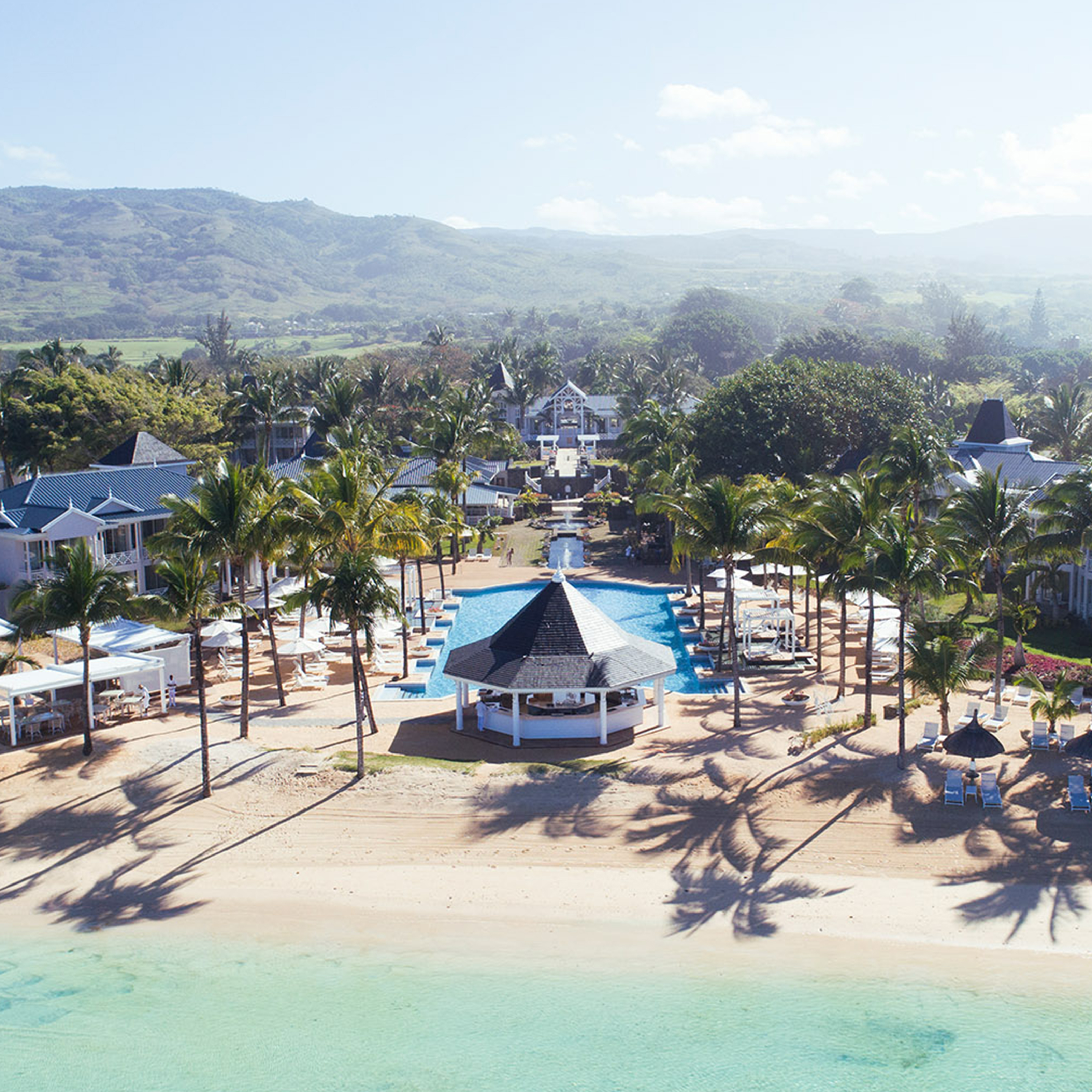 Luftaufnahme eines Strandhotels mit Palmen, Pool, Pavillon und Bergen im Hintergrund. Heller Sandstrand und klares Wasser.