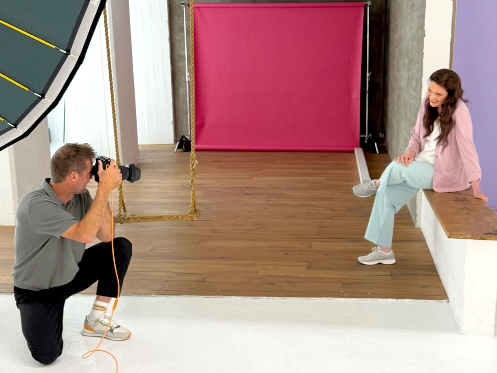 Ein Fotograf kniet und fotografiert eine Frau, die auf einer Bank sitzt. Im Hintergrund sind ein pinker und lila Hintergrund sichtbar. Ein Fotograf kniet und fotografiert eine Frau, die auf einer Bank sitzt. Im Hintergrund sind ein pinker und lila Hintergrund sichtbar.