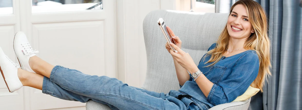 Frau entspannt in einem grauen Sessel, trägt eine blaue Jeanskleidung und weiße Sneaker, während sie ein Smartphone hält. Frau entspannt in einem grauen Sessel, trägt eine blaue Jeanskleidung und weiße Sneaker, während sie ein Smartphone hält.