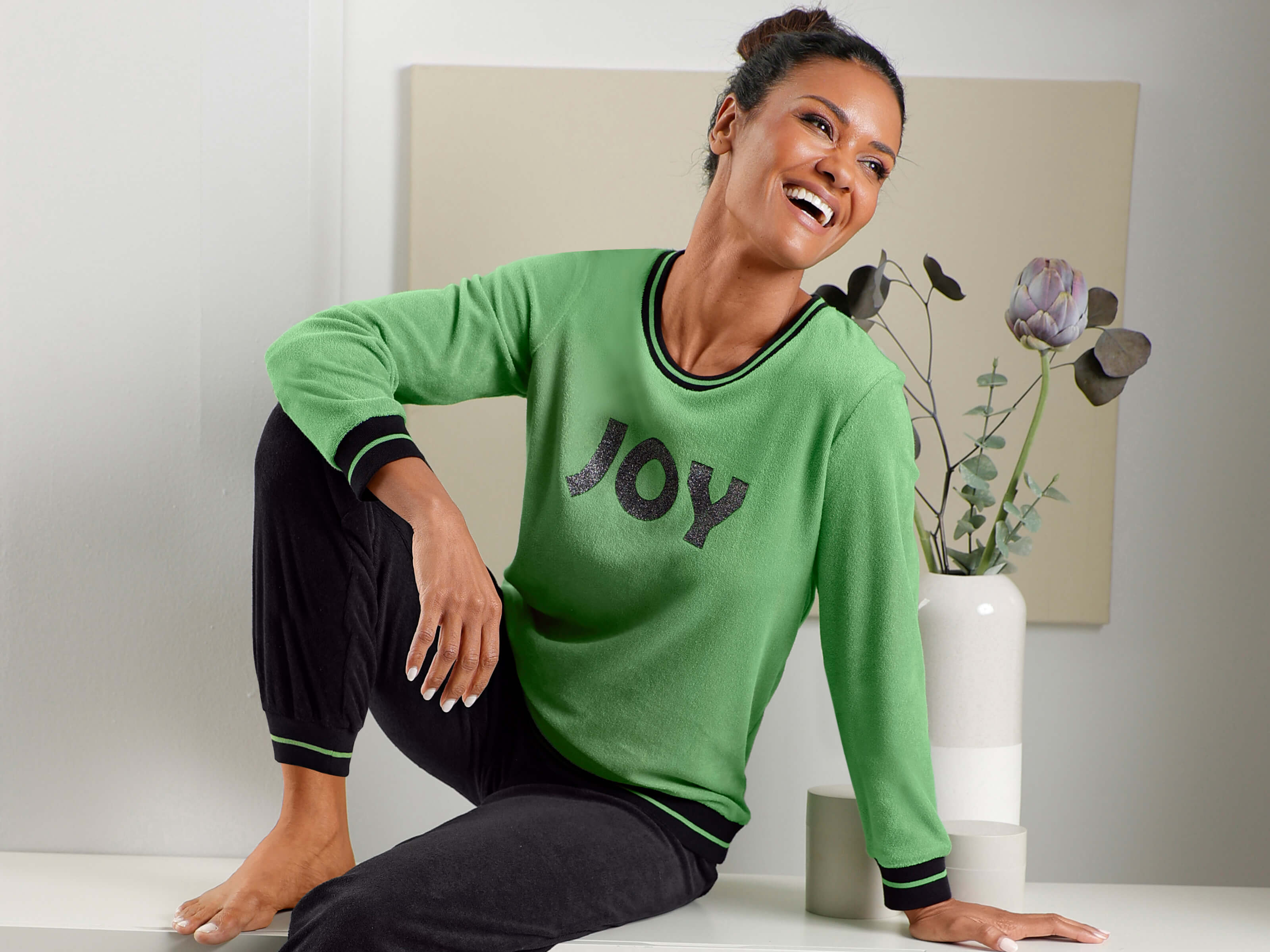 Een model draagt een groene top met het opschrift 'JOY' en een zwarte broek in een lichte ruimte.
