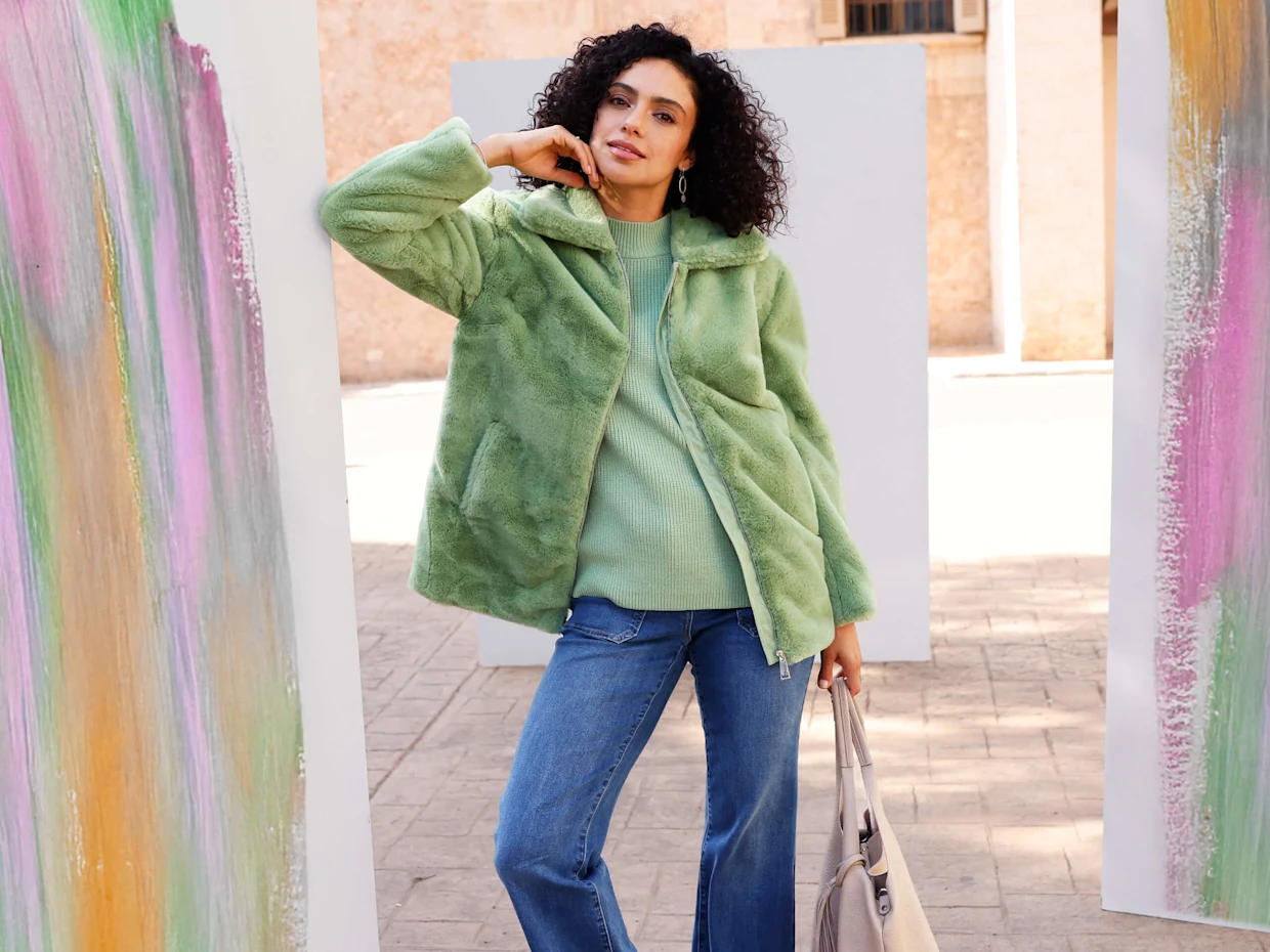 Eine Frau mit dunklen Haaren trägt eine grüne Fleecejacke, einen grünen Pullover und blaue Jeans. Sie steht im Freien, an eine Leinwand mit bunter Farbe gelehnt.