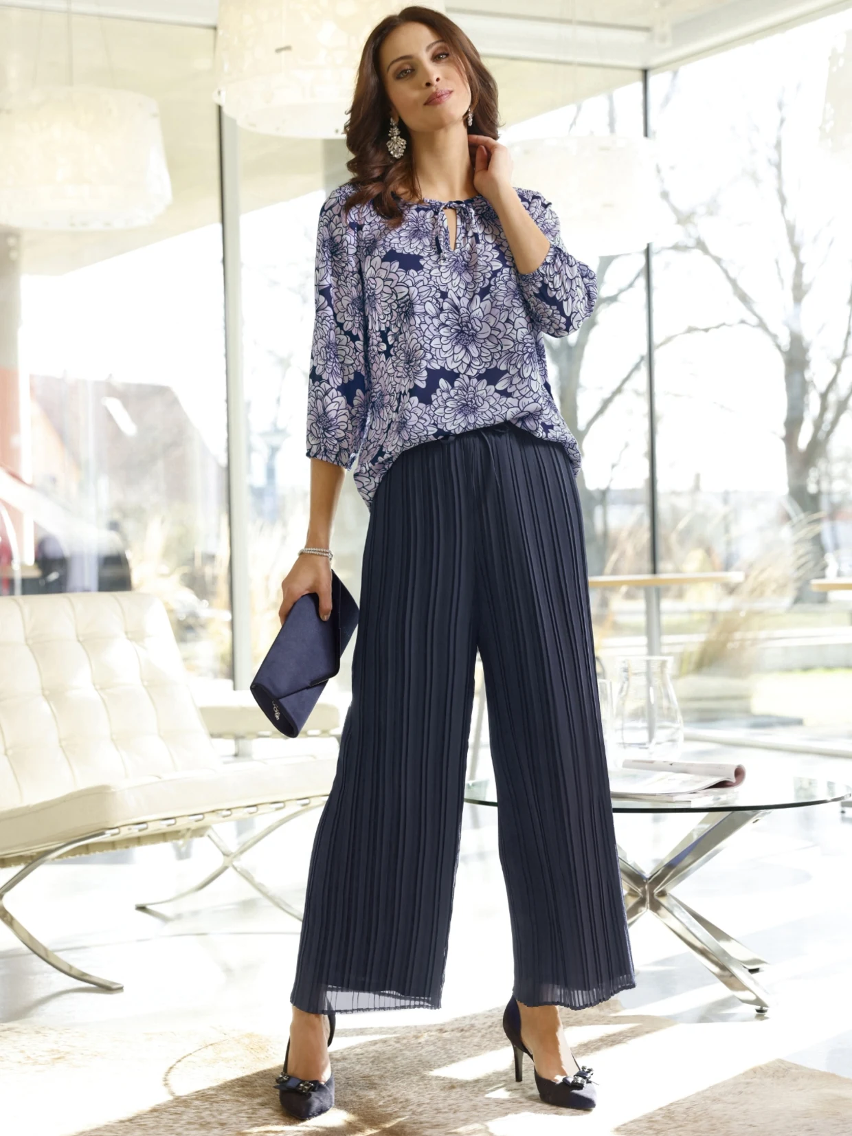 Frau trägt eine elegante blaue Bluse mit floralem Muster und eine weite, plissierte dunkelblaue Hose, kombiniert mit passenden High Heels und einer dunkelblauen Clutch.