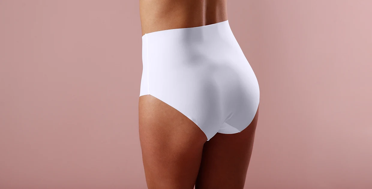 Rückansicht einer Person, die einen weißen, nahtlosen Taillenslip trägt, vor einem rosa Hintergrund.