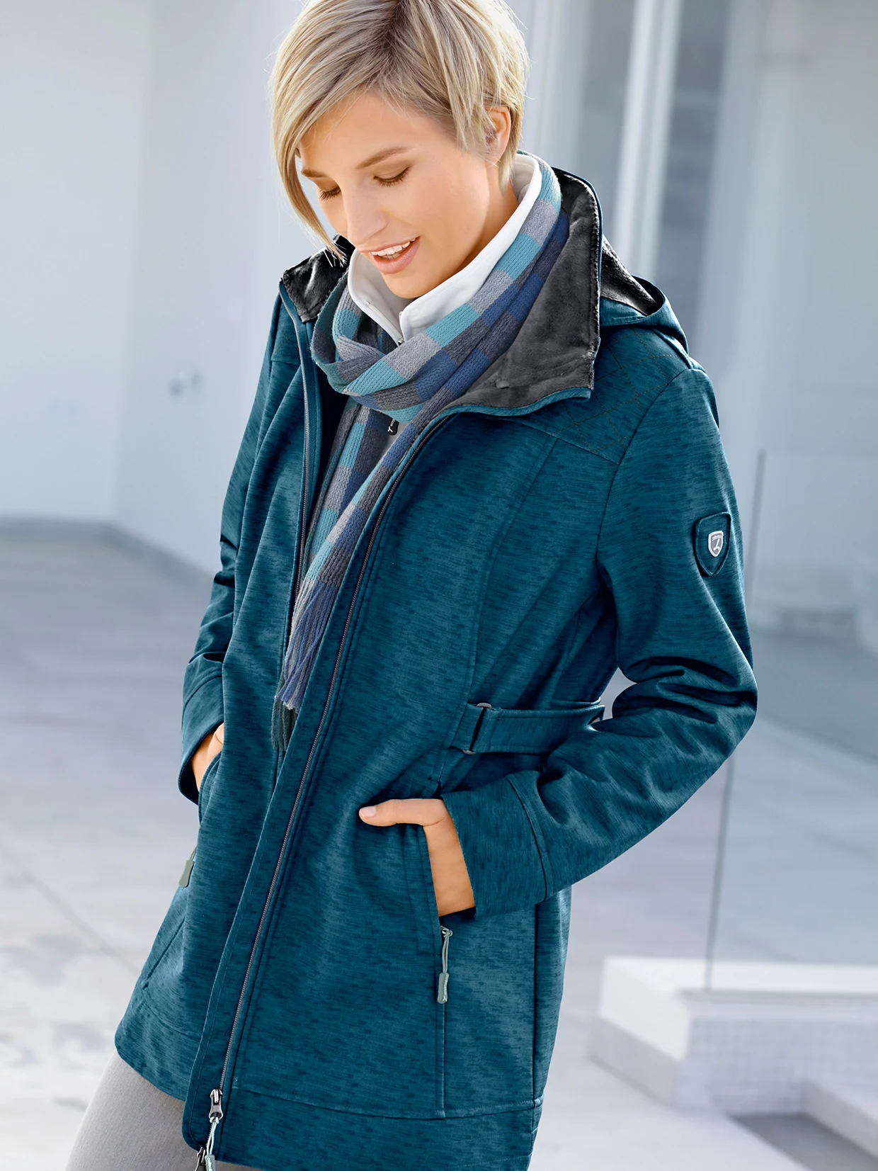 Damenjacke in Petrol mit Reißverschluss, seitlichen Taschen und Stehkragen, kombiniert mit kariertem Schal in Blau-Grau-Tönen.