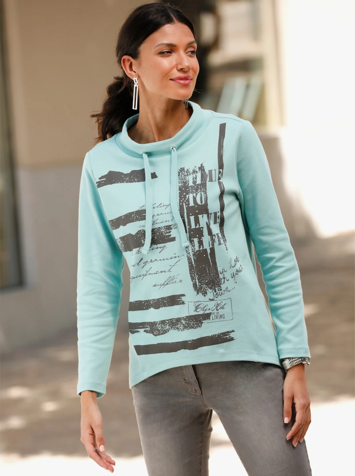 Frau mit türkisfarbenem Sweatshirt mit grauen Glitzersteinchen im Vorderteil und grauer Hose. Frau mit türkisfarbenem Sweatshirt mit grauen Glitzersteinchen im Vorderteil und grauer Hose.
