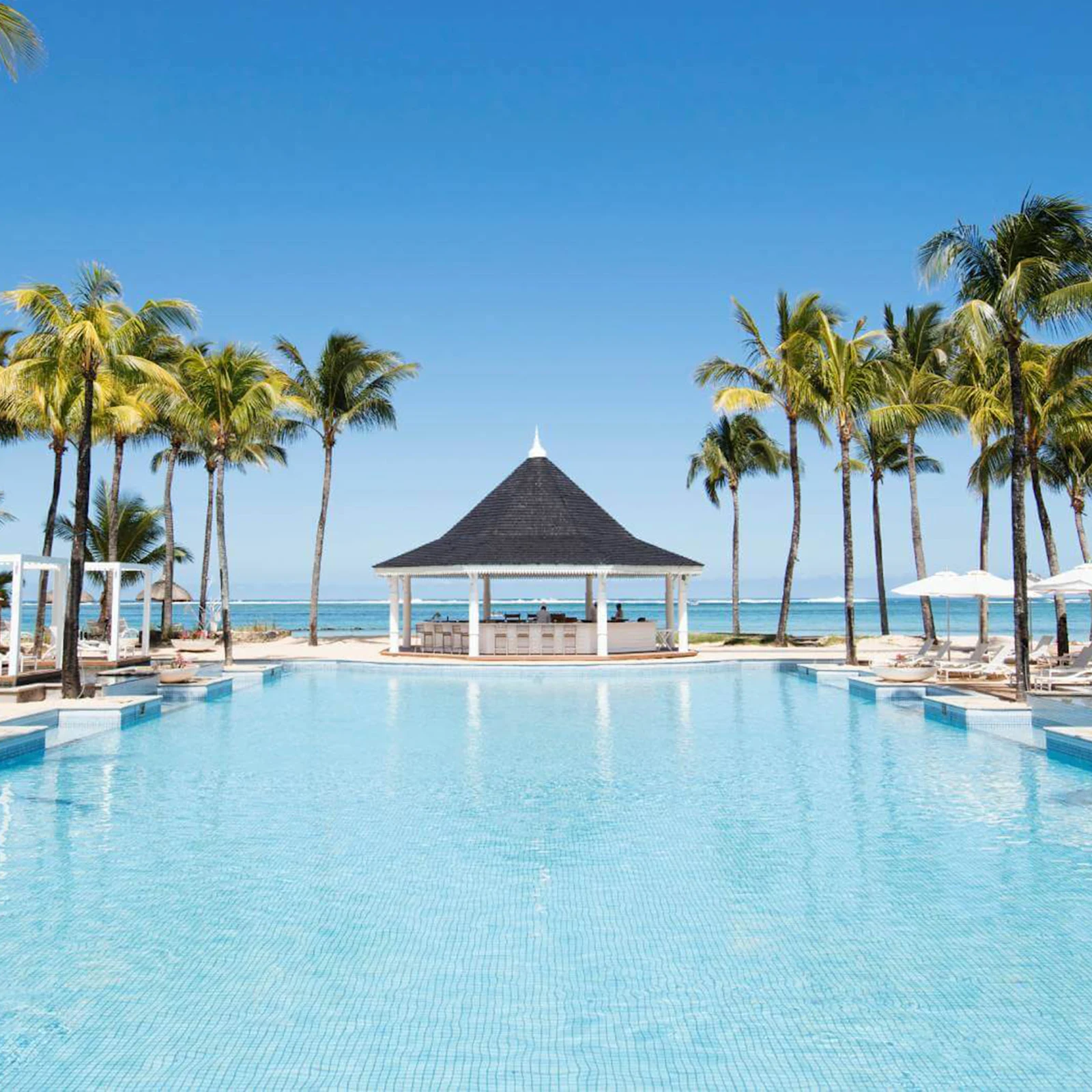 Großer Pool mit klarem Wasser, umgeben von Palmen und weißen Pavillons. Im Hintergrund Meer und blauer Himmel. Großer Pool mit klarem Wasser, umgeben von Palmen und weißen Pavillons. Im Hintergrund Meer und blauer Himmel.