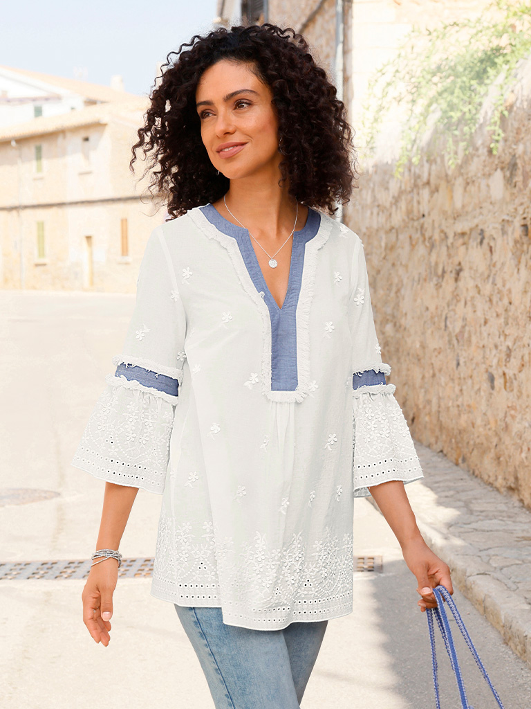 Ein Model trägt eine weiße Tunika mit blauen Details am Ausschnitt und an den Ärmeln, kombiniert mit hellblauen Jeans, stehend im Freien.