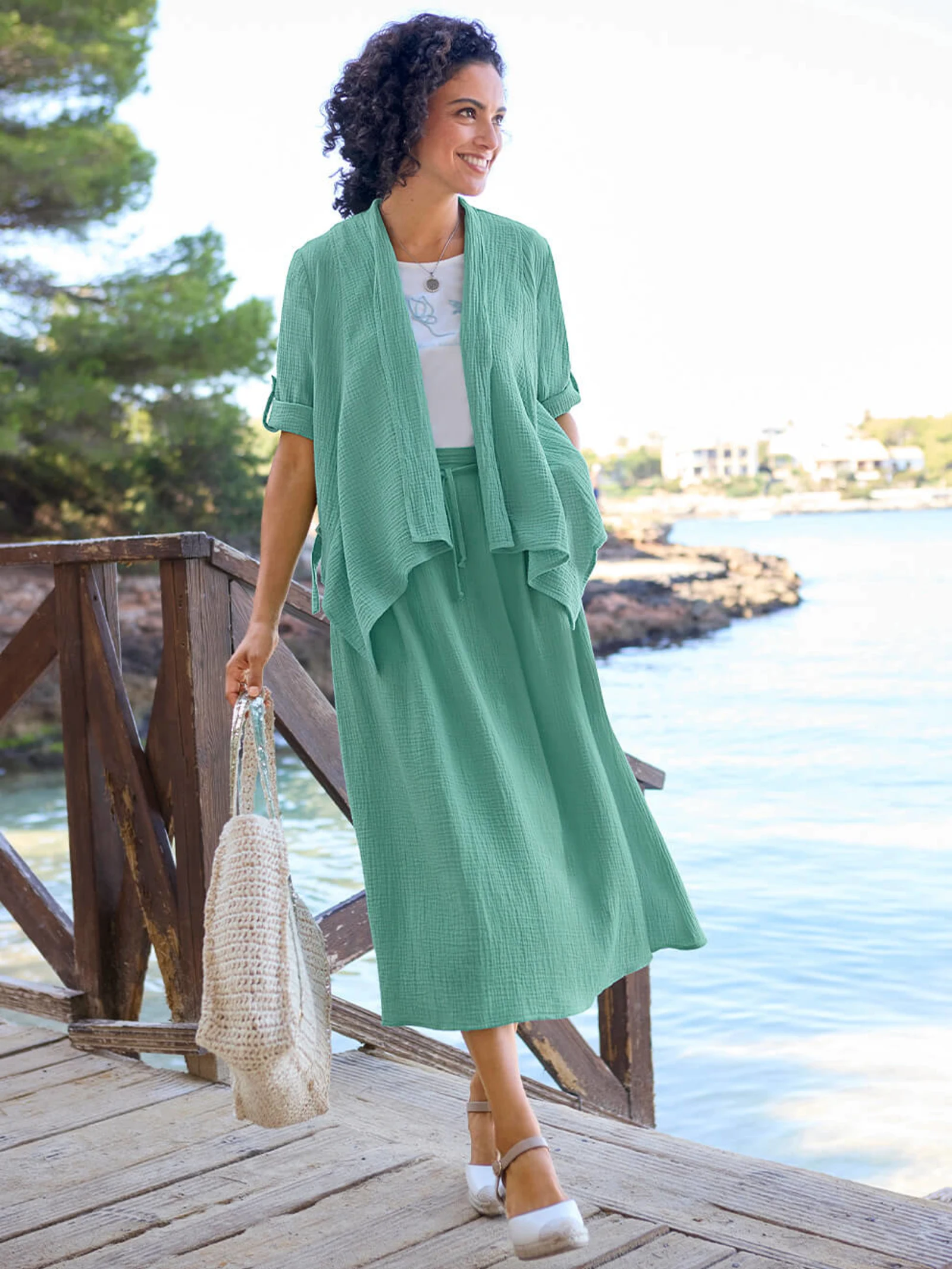 Ein Model trägt eine salbeifarbene Jacke, ein weißes Shirt und einen salbeifarbenen Schlupfrock in Musselin-Qualität, dazu weiße Schuhe und eine gehäkelte Tasche, steht auf einem Steg am Wasser.