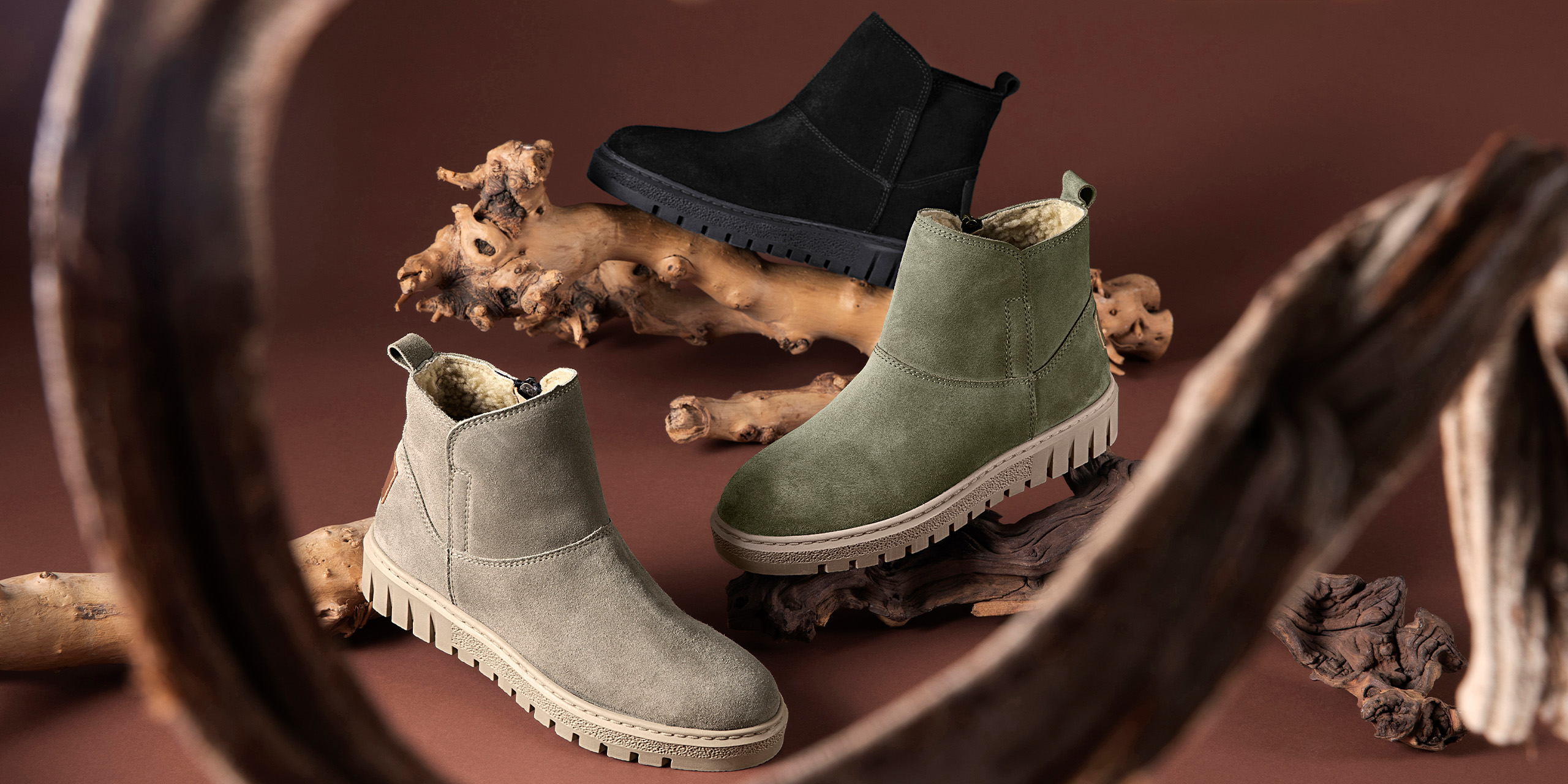 Drei Paar gefütterte Stiefeletten in Schwarz, Olivgrün und Beige auf braunem Hintergrund mit dekorativen Holzstücken.