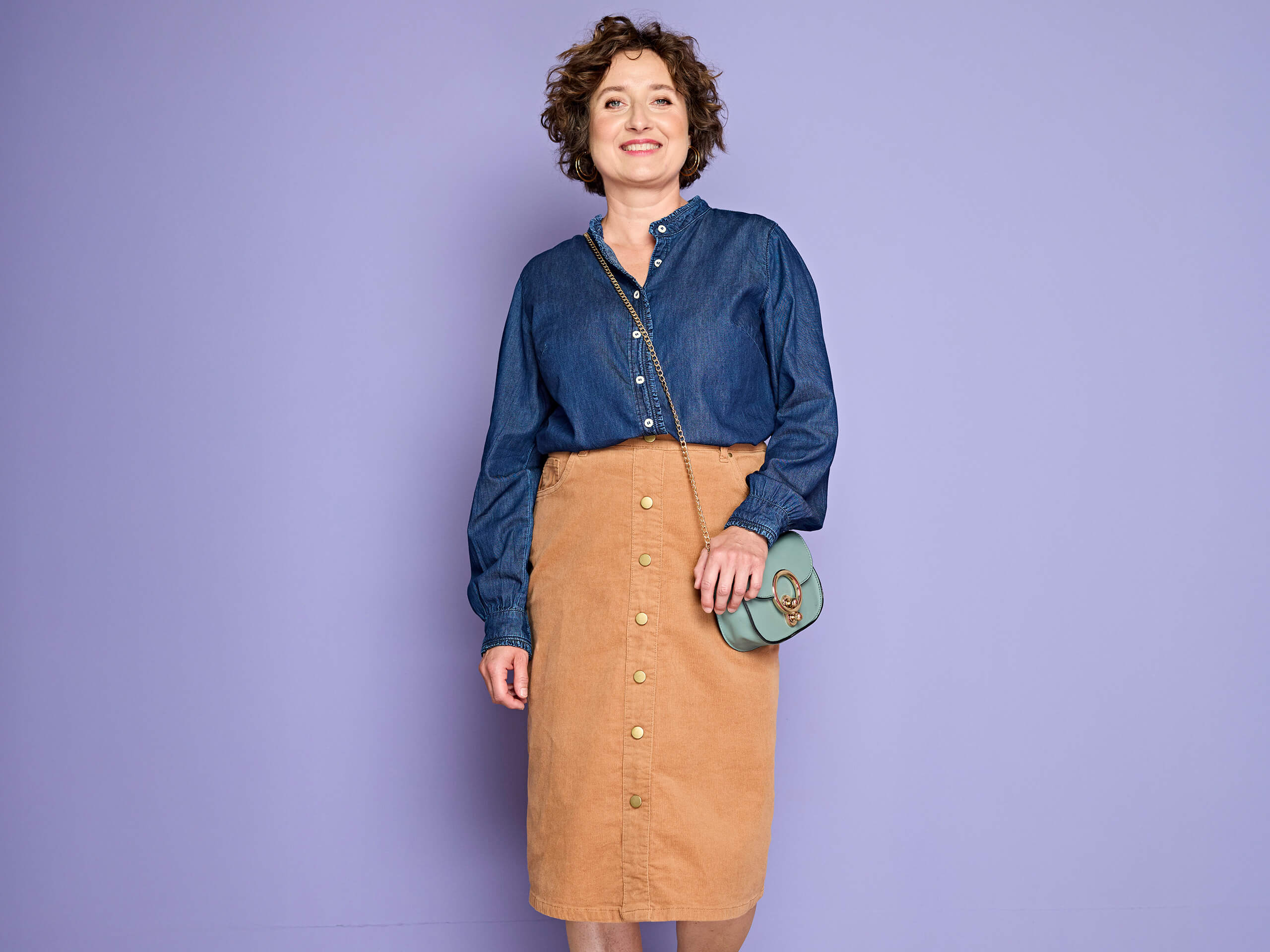 Ein Model trägt eine blaue Jeansbluse, einen braunen Cordrock mit Knöpfen und eine grüne Umhängetasche, vor lila Hintergrund.