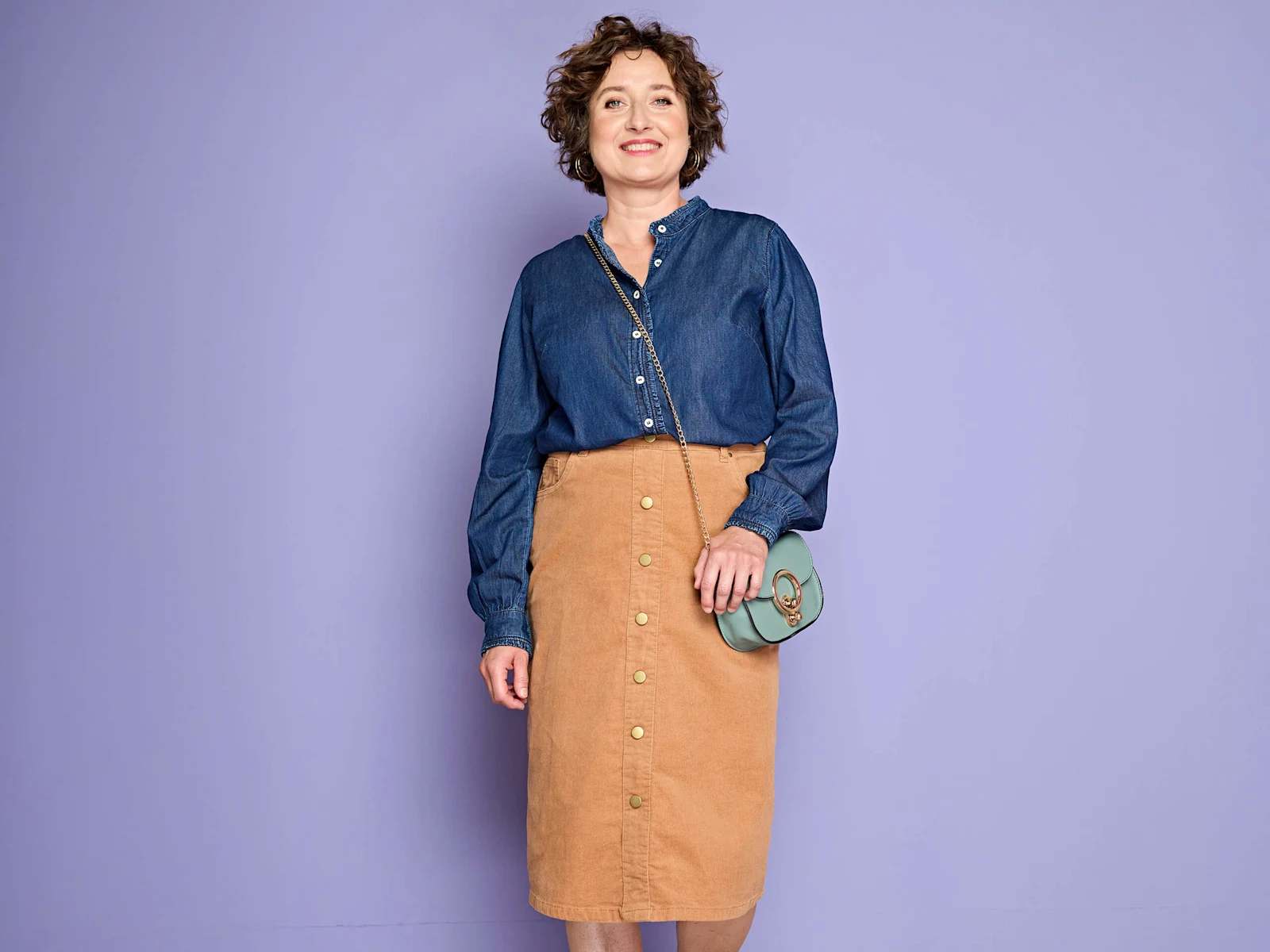Ein Model trägt eine blaue Jeansbluse, einen braunen Cordrock mit Knöpfen und eine grüne Umhängetasche, vor lila Hintergrund.