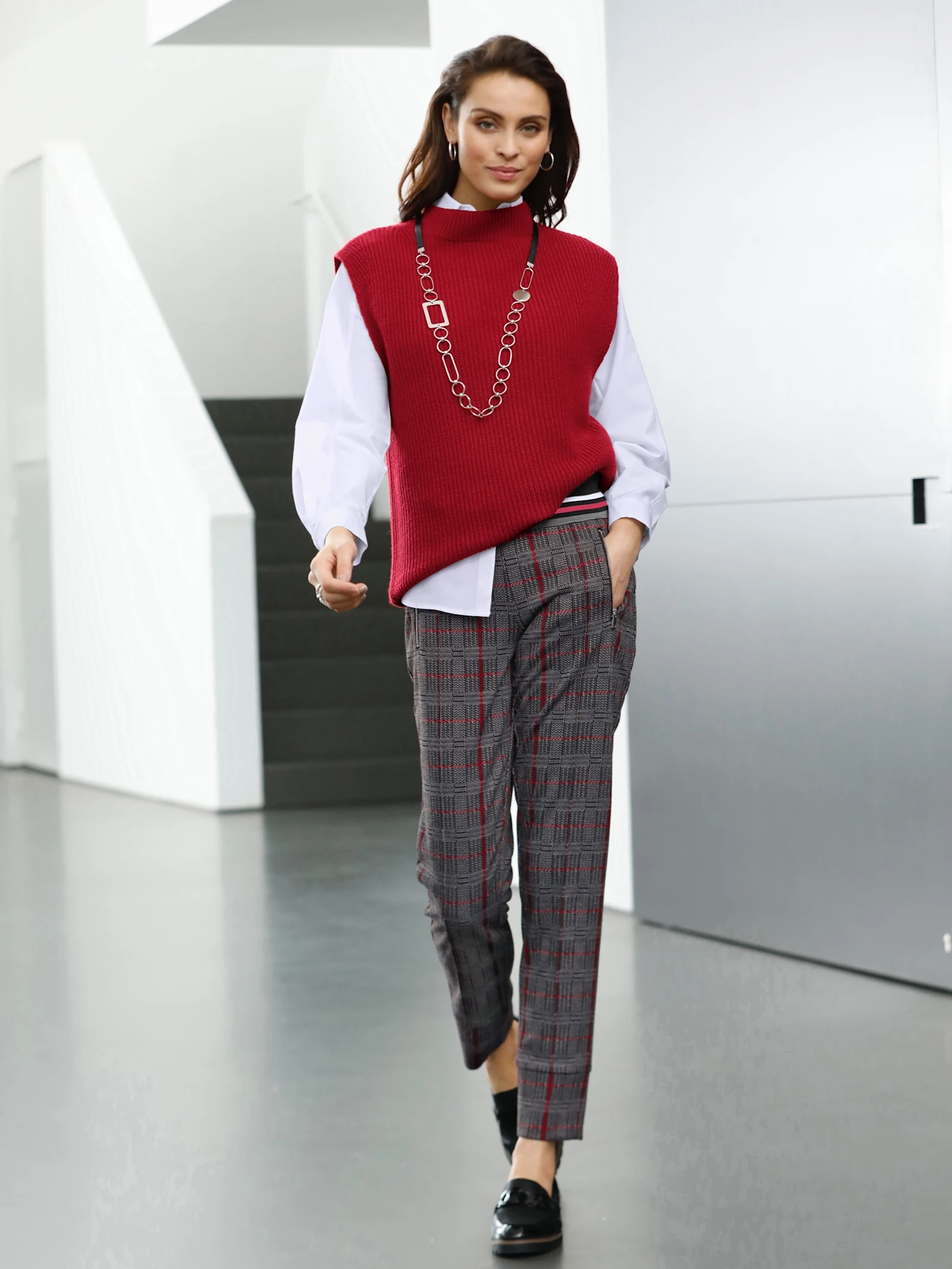 Frau in eleganter Kleidung: Roter Strickpullunder, weiße Bluse, karierte Hose und schwarze Loafer, stilvoll kombiniert in modernem Ambiente. Frau in eleganter Kleidung: Roter Strickpullunder, weiße Bluse, karierte Hose und schwarze Loafer, stilvoll kombiniert in modernem Ambiente.